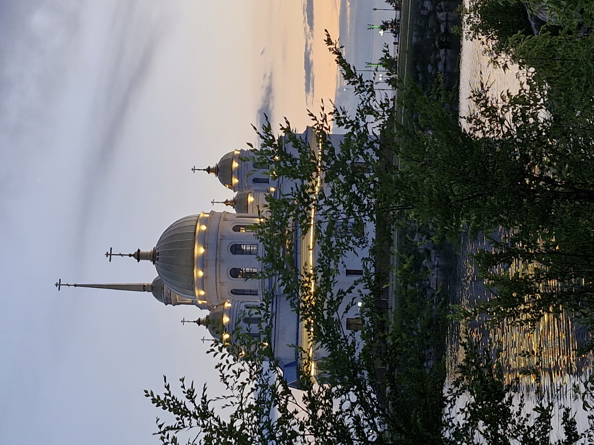 The image depicts the Assumption Cathedral in Kokshetau, Kazakhstan, during twilight. The white cathedral features multiple golden domes topped with crosses, and decorative lights illuminate its exterior. A tall, slender spire rises from behind the main dome. In the foreground, lush green trees with detailed leaves obscure parts of the cathedral and the water. The water, likely a lake or river, reflects the lights from the cathedral and the sky, creating shimmering streaks of light on its surface. The sky is a blend of soft blues and oranges, with wispy clouds indicative of dusk. In the background, a promenade with lampposts and what appear to be benches is visible along the water's edge. The scene is peaceful and serene, capturing the architectural beauty of the cathedral against a natural backdrop at the end of the day.