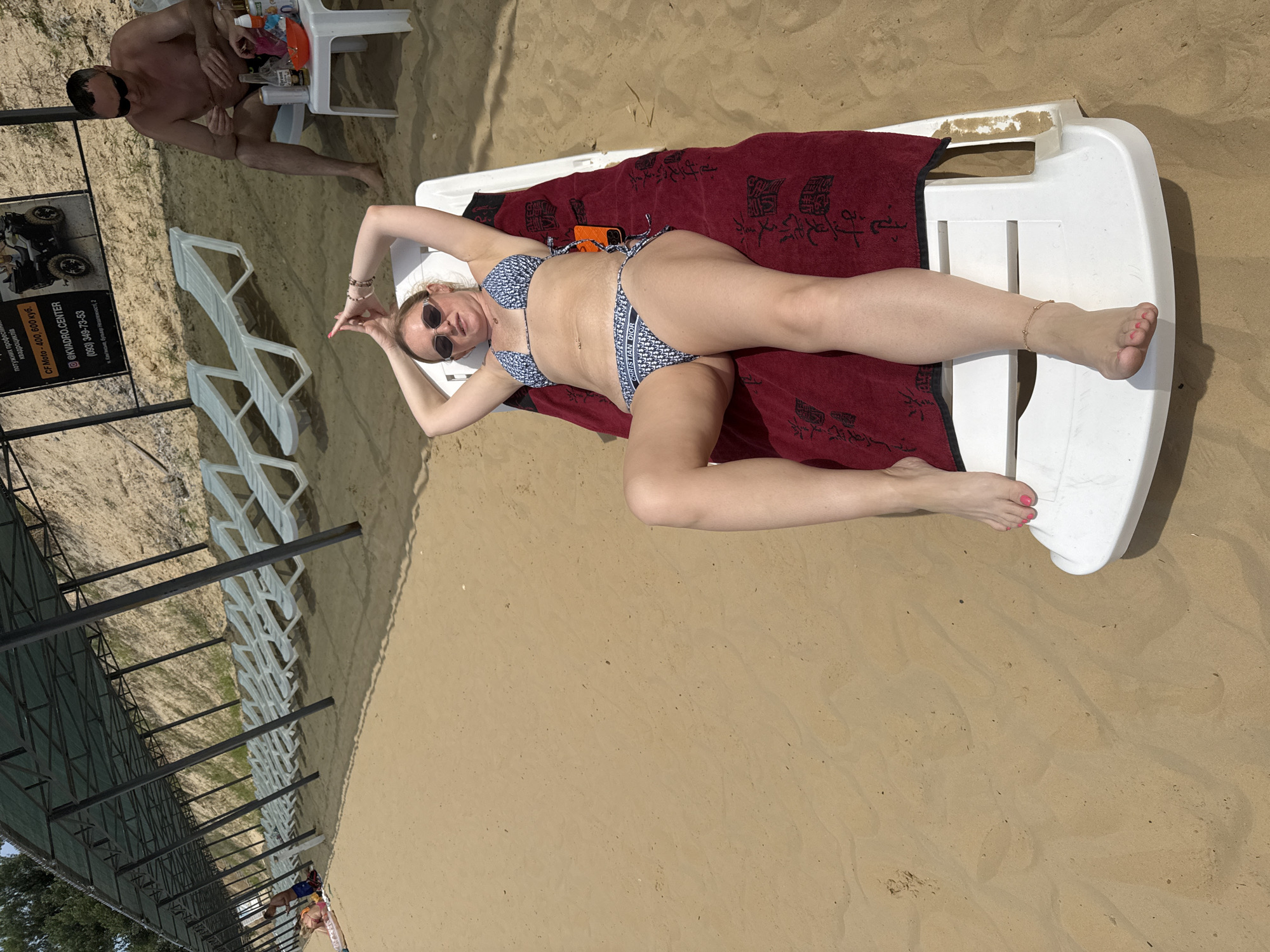 An overhead shot shows a woman lying on a white chaise lounge on a sandy beach. She is wearing a blue and white patterned bikini and sunglasses. Her arms are raised above her head, and she is looking at the camera. A red towel with Asian characters is placed on the chaise lounge beneath her. To the right of the chaise lounge, a man in dark shorts and sunglasses sits on a white plastic chair, holding a bottle and an orange bowl. In the background, rows of empty white lounge chairs are set up under a shaded canopy structure. The beach is made of light brown sand, and in the far background, a sandy slope rises. A sign advertising "CF Moto - 400, 600 kub." and a website "@KVADRO.CENTER" is visible on the canopy structure. The location is Yelyzavetivka, Ukraine.