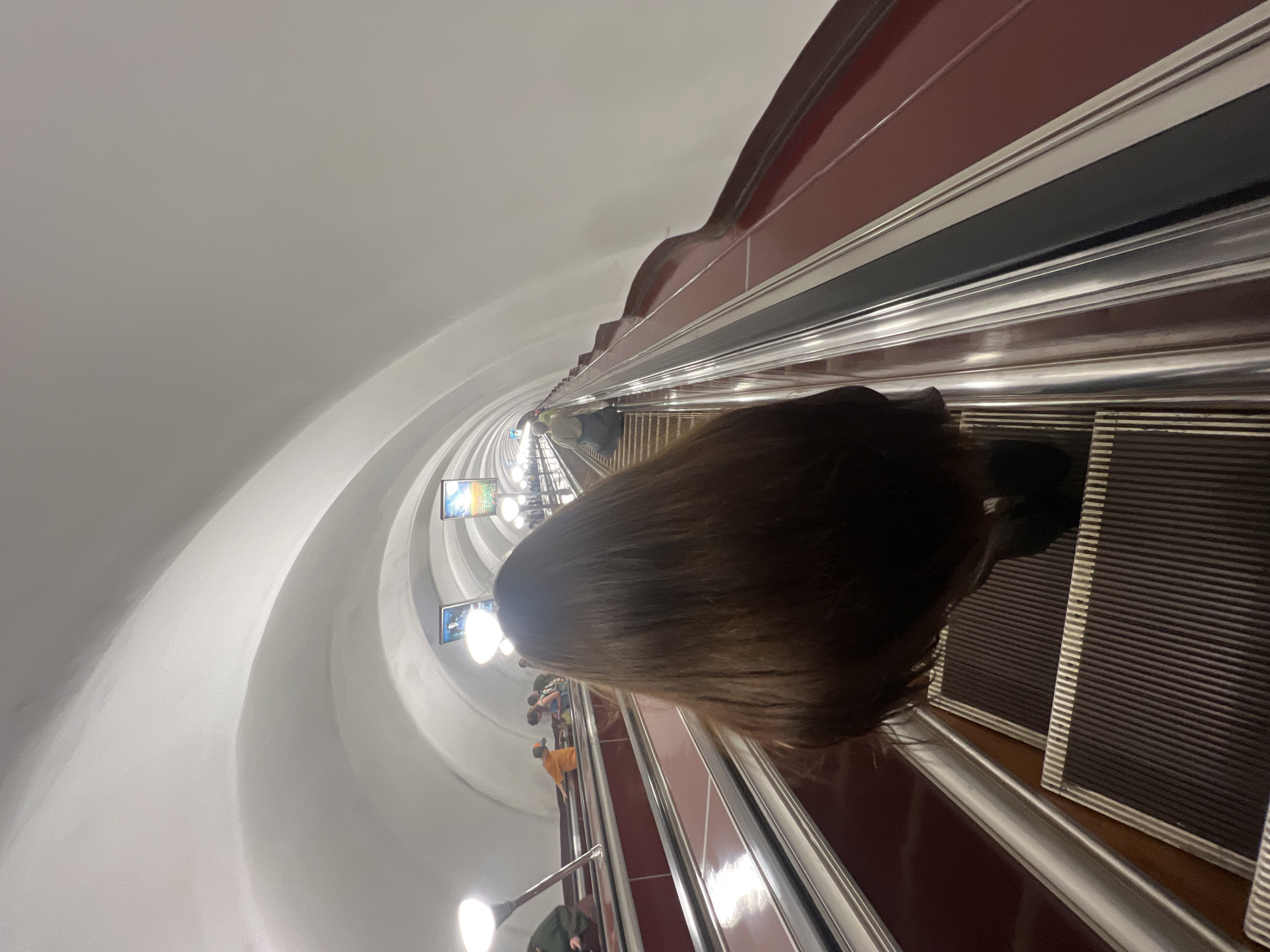The image shows a person with long, dark brown hair descending an escalator in the Saint Petersburg Metro. The escalator has a red-colored side panel and silver-colored handrails. The ceiling of the metro station is white and arched, with lighting fixtures and framed posters visible along the tunnel. Other passengers are seen on the escalator and platform in the background, some walking and others standing. The scene appears to be an interior view of a metro station, characterized by its subterranean architecture and functional design.