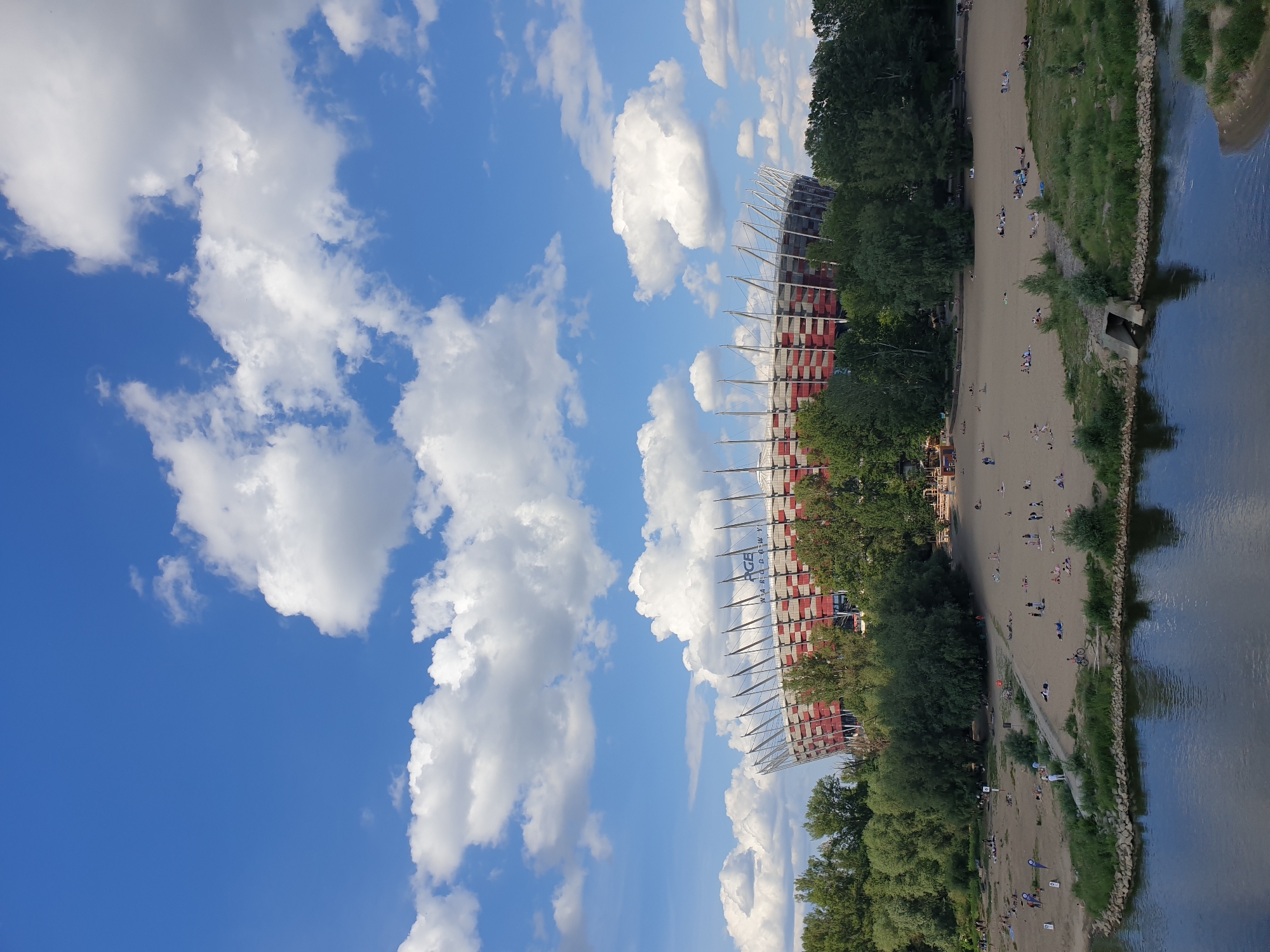 The image captures a wide-angle view of the PGE Narodowy stadium in Warsaw, Poland, overlooking the Vistula River. The sky is bright blue, filled with numerous fluffy white cumulus clouds of varying sizes.
In the middle-ground, the PGE Narodowy stadium is prominent, partially obscured by a line of lush green trees. The stadium's exterior features a distinctive architectural design with alternating red and white panels and a series of white, upward-slanting structural supports. The words "PGE NARODOWY" are visible in dark blue lettering on a light background near the top of the stadium's facade.
Between the stadium and the river, a wide, light brown sandy beach stretches along the riverbank. Approximately 50 to 70 individuals and small groups are dispersed across this sandy area, engaged in leisure activities such as sunbathing (many lying down on towels or directly on the sand), sitting, and walking. Some people are seen conversing or observing the surroundings. A narrow path of lighter sand runs through the beach. A single bicycle is visible on the sand near the water's edge on the right. Small patches of green grass and bushes are interspersed with the sand, particularly closer to the river and at the edges of the beach area.
The foreground is dominated by the dark blue-grey water of the Vistula River, reflecting patches of the sky. The river's surface is relatively calm with minor ripples. On the right side of the riverbank in the immediate foreground, a concrete or stone-lined culvert empties into the water. The riverbank itself is covered with some rocks and green vegetation. The scene is well-lit under clear daylight conditions.