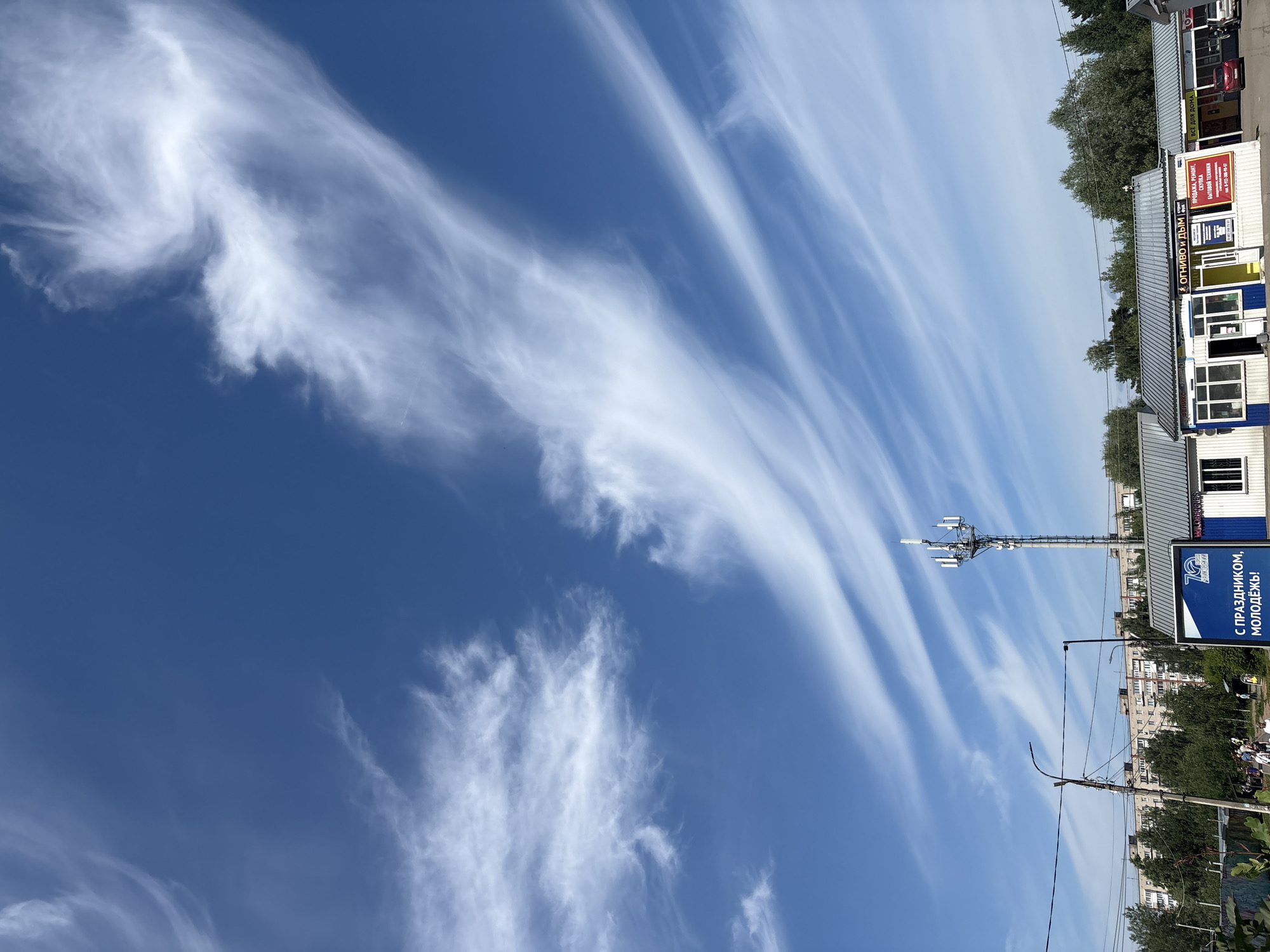 The image depicts a street scene in a city. The sky is a vibrant blue with wispy white clouds. In the foreground, there is a telecommunications tower. To the right of the tower, there are several small buildings with storefronts. One storefront has a red and white sign with Russian text. Further to the left, there are apartment buildings and trees. A few people are visible walking on the sidewalk in front of the apartment buildings. The overall scene suggests a sunny day in an urban environment.