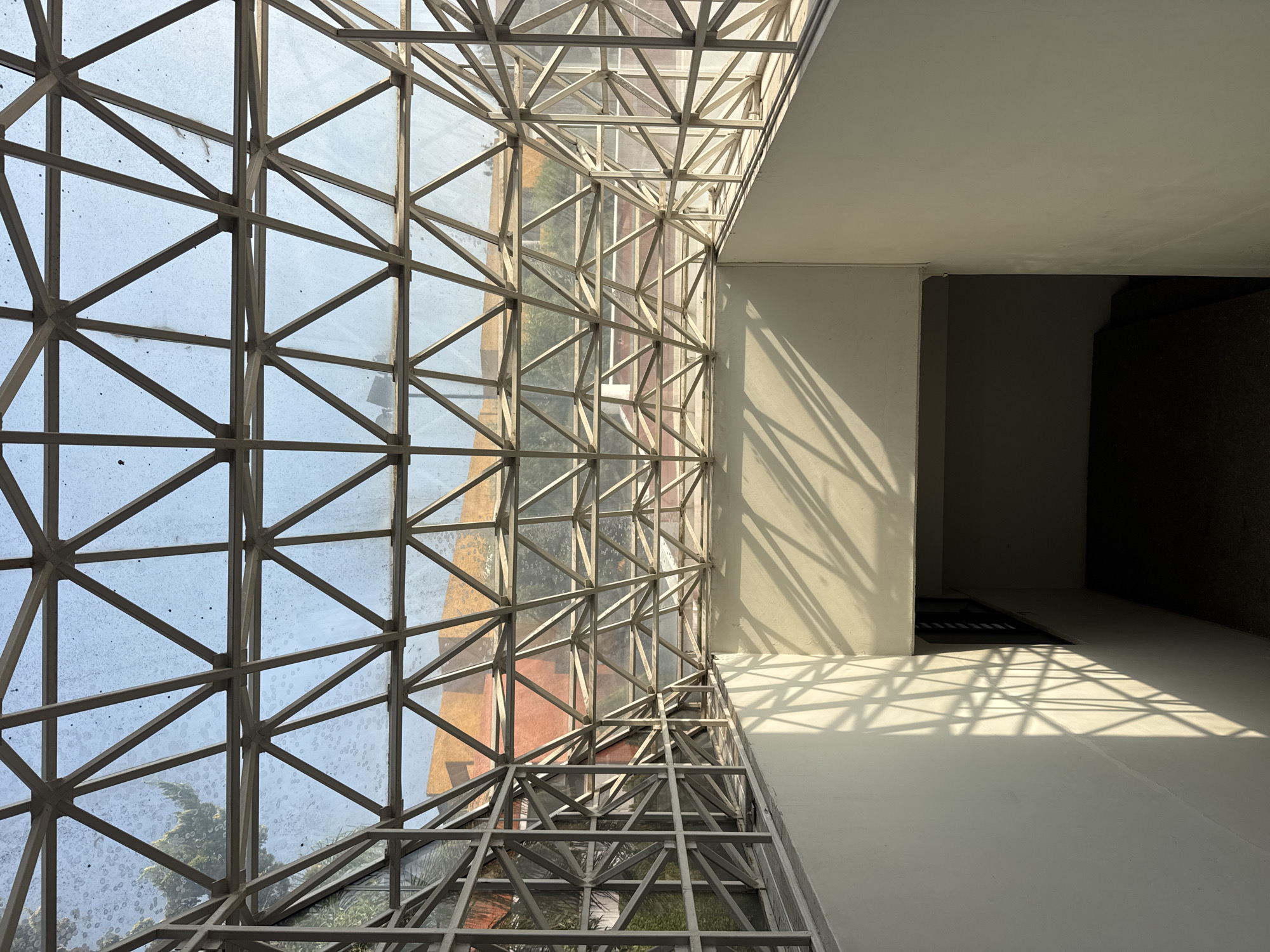 The image presents a striking architectural interior, likely a corridor or an atrium, characterized by a large, geometric metal and glass structure dominating the left side of the frame. This structure, possibly a skylight or a multi-story window wall, is composed of numerous interconnected metal beams forming a complex grid of triangles and rectangles, enclosing panes of glass. The glass appears somewhat dusty or speckled, hinting at exposure to the elements.
Through the glass, glimpses of the exterior environment are visible, including green foliage from trees and parts of an adjacent building featuring warm, possibly terracotta-colored walls and subtle patterns or textures.
The right side of the image reveals a clean, light-colored interior wall and ceiling, creating a stark contrast with the elaborate glass structure. Strong, directional sunlight pierces through the metal framework, casting dramatic, sharp-edged geometric shadows across these interior surfaces. These intricate patterns of light and shadow extend onto what appears to be a light-colored floor, suggesting a bright and sunny day, likely during mid-morning or afternoon.
Further into the interior space, towards the lower right and in darker recesses, a deep shadow obscures parts of the building's design, possibly indicating a doorway, a hidden passageway, or a stairwell. No individuals or visible text are present in the image. The scene focuses on the interplay of light, shadow, and modern architectural elements within a setting in San Andrés Cholula, Mexico.