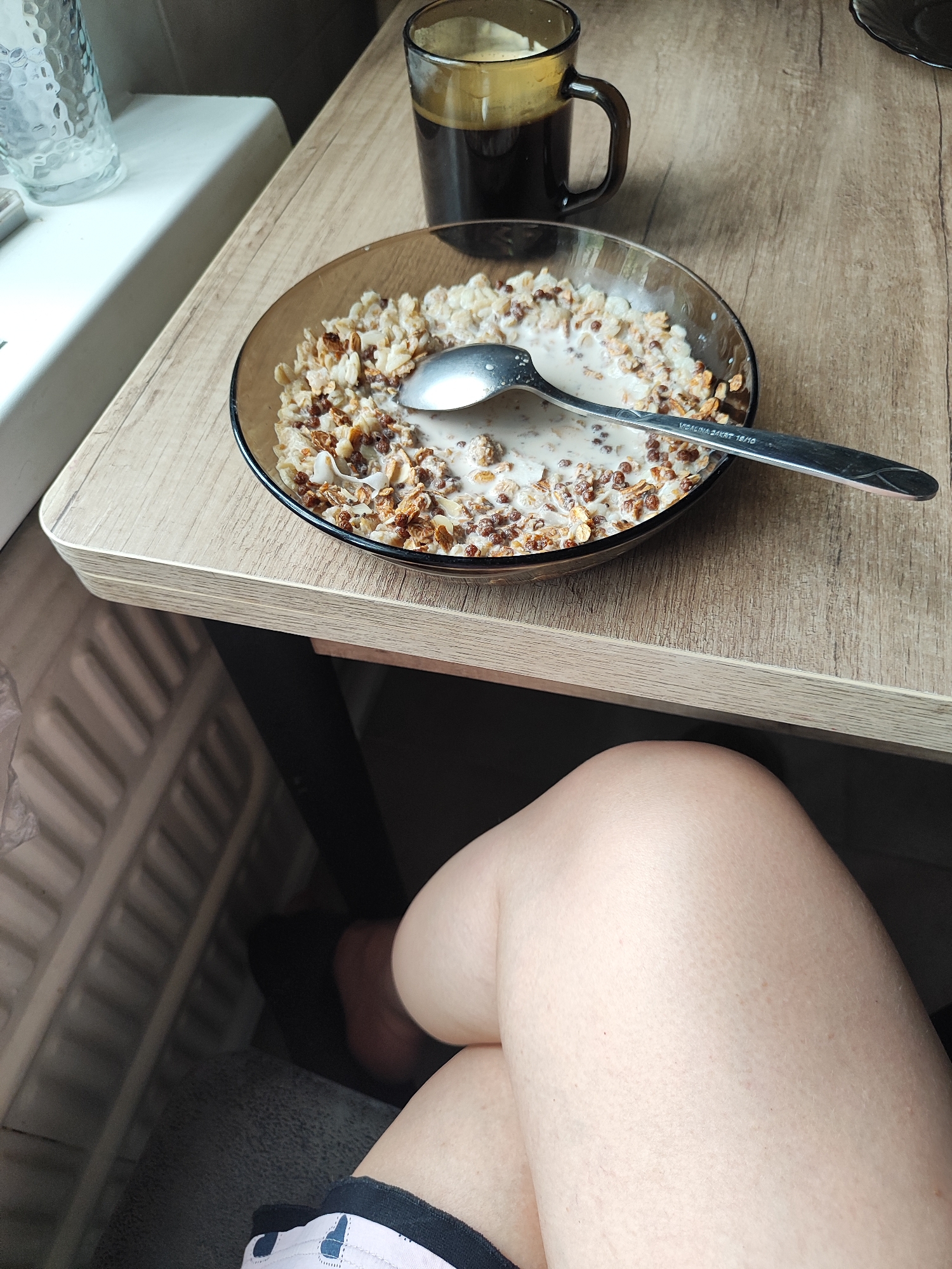 A close-up overhead shot shows a bowl of cereal with milk and a spoon on a wooden table. Next to the bowl is a glass mug of dark coffee. The scene appears to be set indoors, possibly on a windowsill, as a textured white ledge is visible in the background. The lower part of the image shows a person's legs and thighs, indicating their presence in the frame. The lighting suggests it is daytime. There is no identifiable information in the image that places it specifically in Almaty, Kazakhstan.