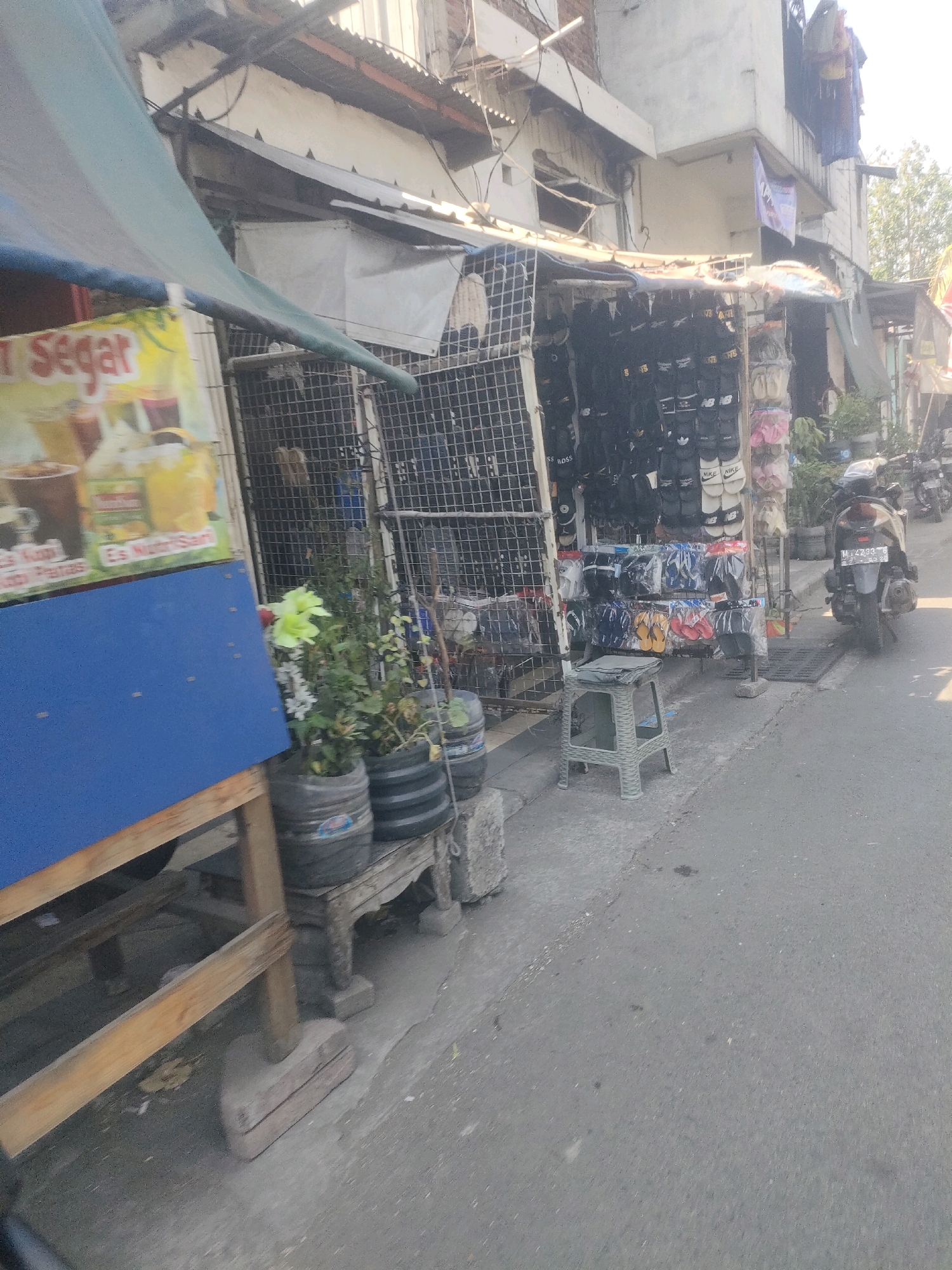 This image depicts a street scene in Surabaya, Indonesia, likely a commercial area. In the foreground, a blue board is visible, part of a stall. To its right, a selection of footwear, predominantly flip-flops and sandals, is displayed on racks. Behind the footwear, a metal fence encloses more of the stall. Further to the right, a motorcycle is parked on the asphalt road. The background shows a two-story building with a corrugated metal roof, characteristic of urban housing or small businesses in the region. Laundry is hanging from the upper floor of the building. The overall environment appears to be a busy street with informal vendors.