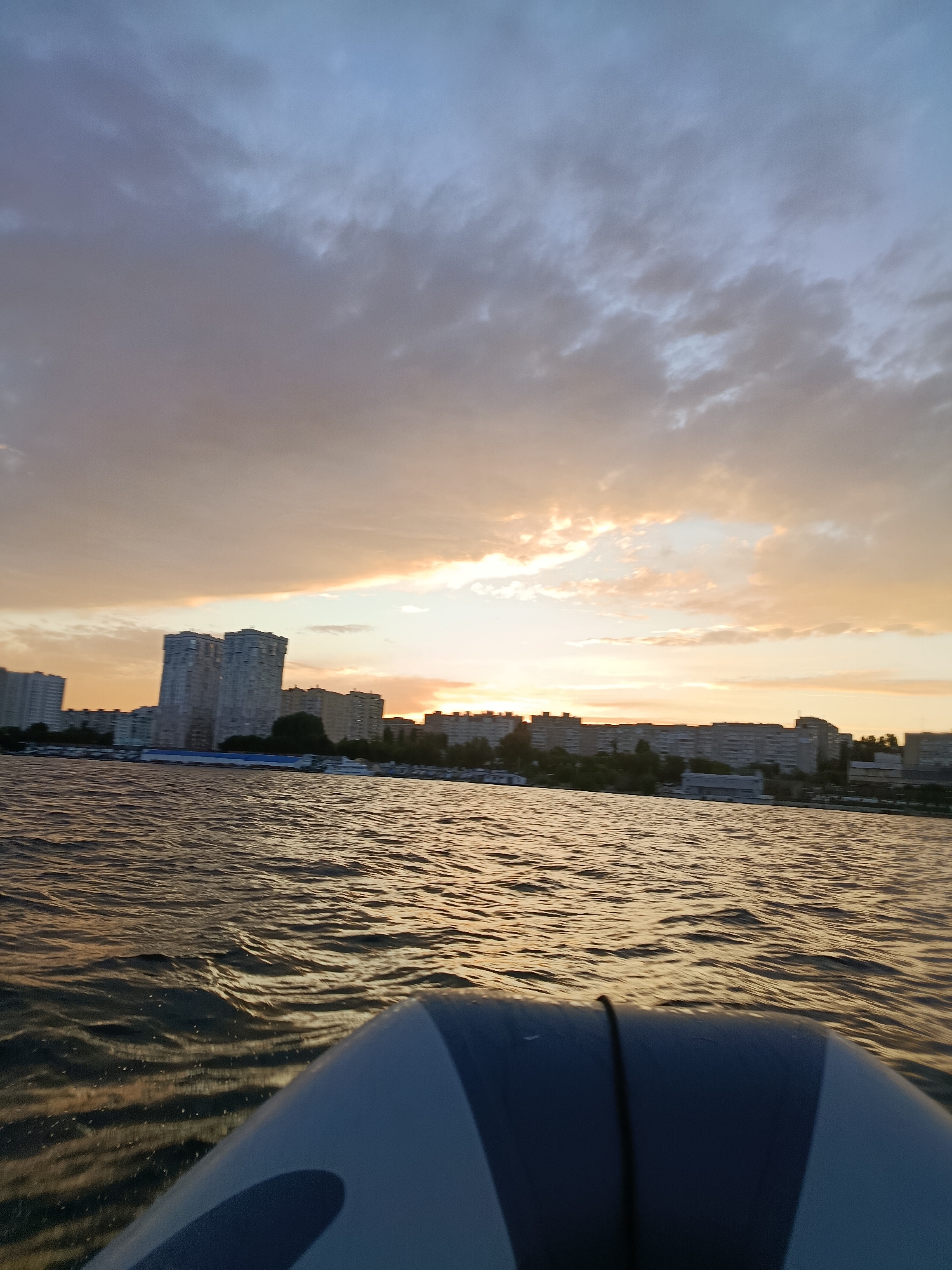 An outdoor, low-angle, wide shot captures a cityscape at sunset viewed from the front of a boat. The foreground is dominated by the bow of an inflatable boat, gray and white, with a black rope running through it. The water is choppy and reflects the golden hues of the sunset. In the background, a long line of apartment buildings stretches across the horizon. The sky is filled with dramatic clouds, tinged with orange and purple from the setting sun. A few tall buildings stand out amongst the lower structures. The scene appears to be on a river, likely the Volga River, given the context of Saratov, Russia. There are no people visible in the image. The overall impression is one of a peaceful, serene evening on the water with a urban backdrop.