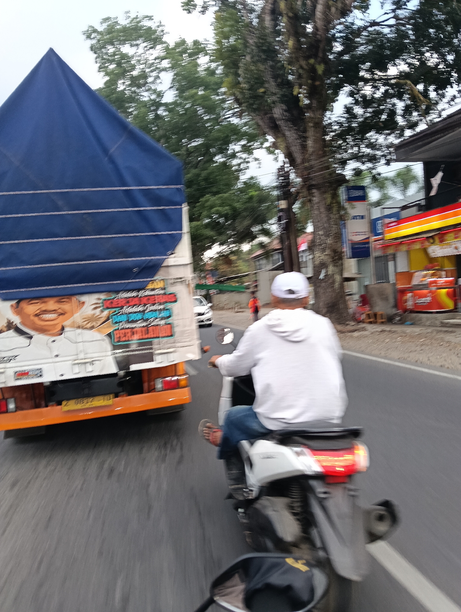 In Cintaraja, Indonesia, a person on a white scooter, wearing a white hooded sweatshirt and blue jeans, is captured in motion on a paved road. The scooter's taillight is illuminated red. The scooter is positioned behind a truck with a blue tarp covering its cargo. The side of the truck features a large painted image of a smiling man in a white uniform, along with text. The truck's license plate is visible and reads "Z 9617 TD". In the background, a line of buildings with awnings, including one with red and yellow signage, and trees with lush green foliage line the road. A white car is also visible in the distance. The scene suggests a typical day in a bustling Indonesian town.