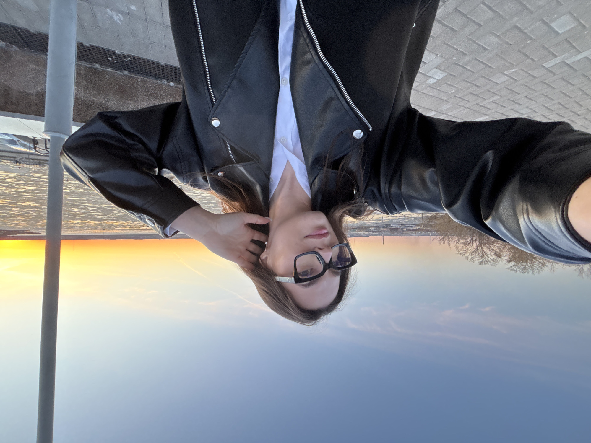 A woman is featured in a selfie, looking directly at the camera with a calm expression. She has long, dark hair and wears black-framed glasses. She is dressed in a white collared shirt, open at the neck, layered under an unzipped black leather jacket with silver hardware accents. Her right hand is gently touching the back of her head or hair.
The setting is outdoors during sunset. To the woman's left, a vibrant sunset illuminates the horizon with bright orange and yellow hues, fading into a pale blue sky above a body of water. Distant, bare trees and faint outlines of structures, possibly part of a city skyline, are visible on the far horizon to her right. The foreground shows a paved area, which appears to be a footpath or embankment, and a light gray vertical pole stands prominently on the far right side of the frame. The overall atmosphere is peaceful, characterized by the soft, warm light of the setting sun. There is no visible text in the image.