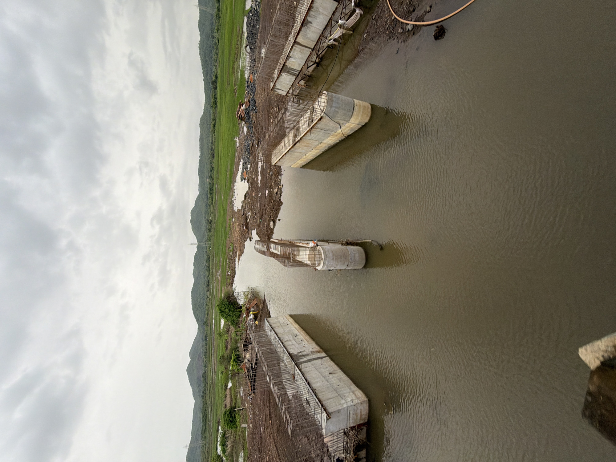 The image depicts a construction site for a bridge in Saphale, India. The scene shows several concrete pillars and beams emerging from murky water, indicating that the bridge is under construction over a river or canal. The surrounding landscape is a mix of undeveloped land with piles of construction materials and lush green fields with rolling hills in the background. The sky is overcast with heavy clouds, suggesting a possibly rainy or humid day. There are no visible people actively working in the immediate foreground of the construction, but a single figure appears to be present near one of the pillars. The overall impression is of a large infrastructure project in progress within a natural, undeveloped setting.