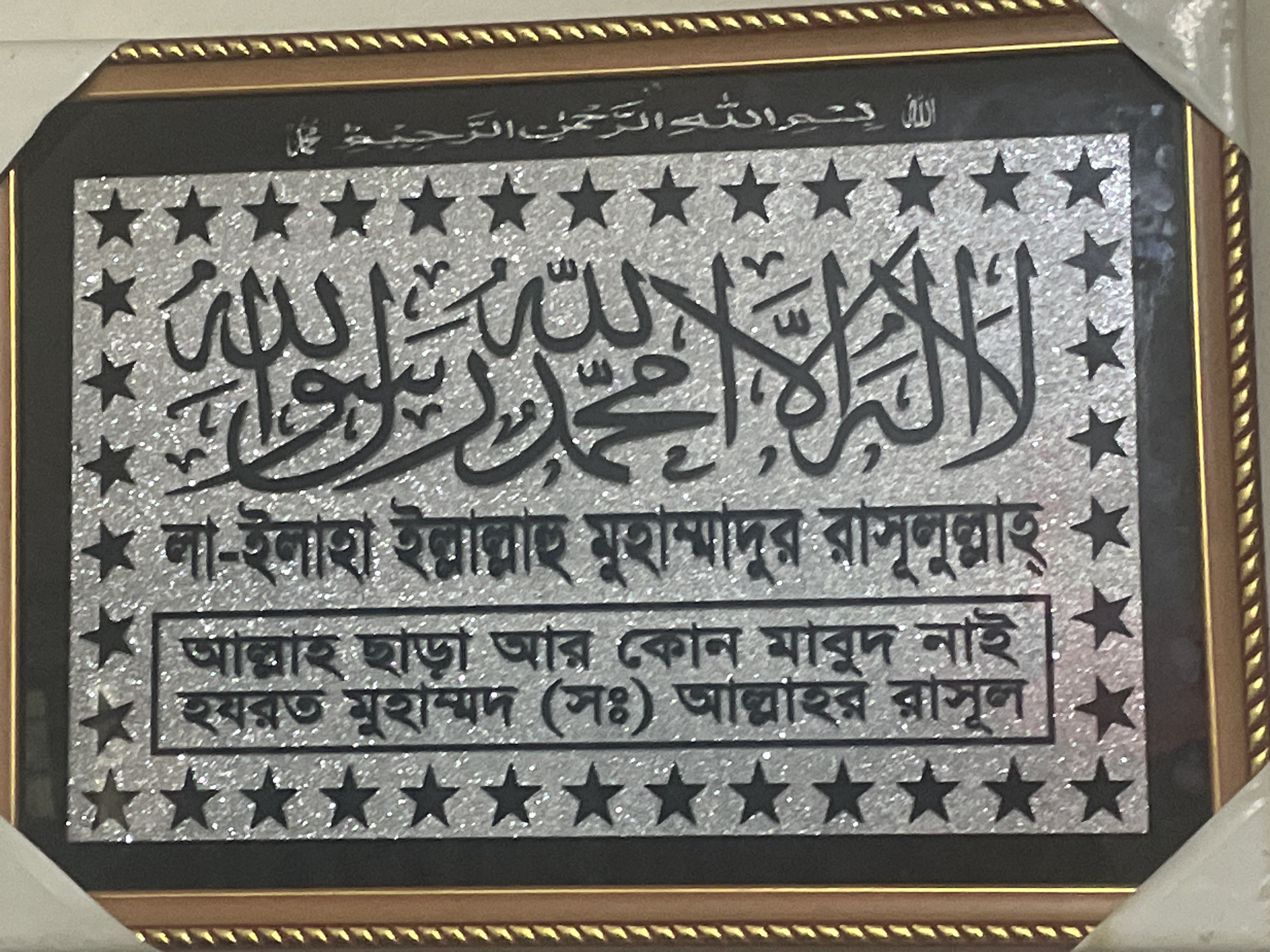 Here's a detailed description of the image:
**General Description:**
The image shows a framed decorative plaque featuring Arabic calligraphy and Bengali script. The piece has a shimmering silver background with black lettering and star motifs.
**Content Details:**
* **Top Line (Arabic):** Displays "Bismillah Al-Rahman Al-Rahim," a common Islamic phrase meaning "In the name of God, the Most Gracious, the Most Merciful."
* **Middle Section (Arabic):** Presents the Shahada, the Islamic declaration of faith: "La Ilaha Illallah Muhammadur Rasulullah" (There is no god but Allah, Muhammad is the messenger of Allah).
* **Lower Section (Bengali):** Contains a translation and explanation of the Shahada in Bengali: "Allah chara ar kono mabud nai; Hazrat Muhammad (SAW) Allah'r Rasul" (There is no god but Allah; Hazrat Muhammad (peace be upon him) is the messenger of Allah).
**Visual Elements:**
* The background has a reflective, glittery silver finish.
* Black stars adorn the top and bottom margins.
* The frame appears to be gold-colored with a textured design.
* The piece overall looks to be a modern interpretation of traditional Islamic art.
**Possible Location:**
The item's style and the presence of Bengali script strongly suggest a location where Bengali speakers are prevalent, such as in Dhaka, Bangladesh.
**Overall Impression:**
The plaque is a decorative item likely intended for display in a home or office, reflecting religious sentiment and artistic expression.