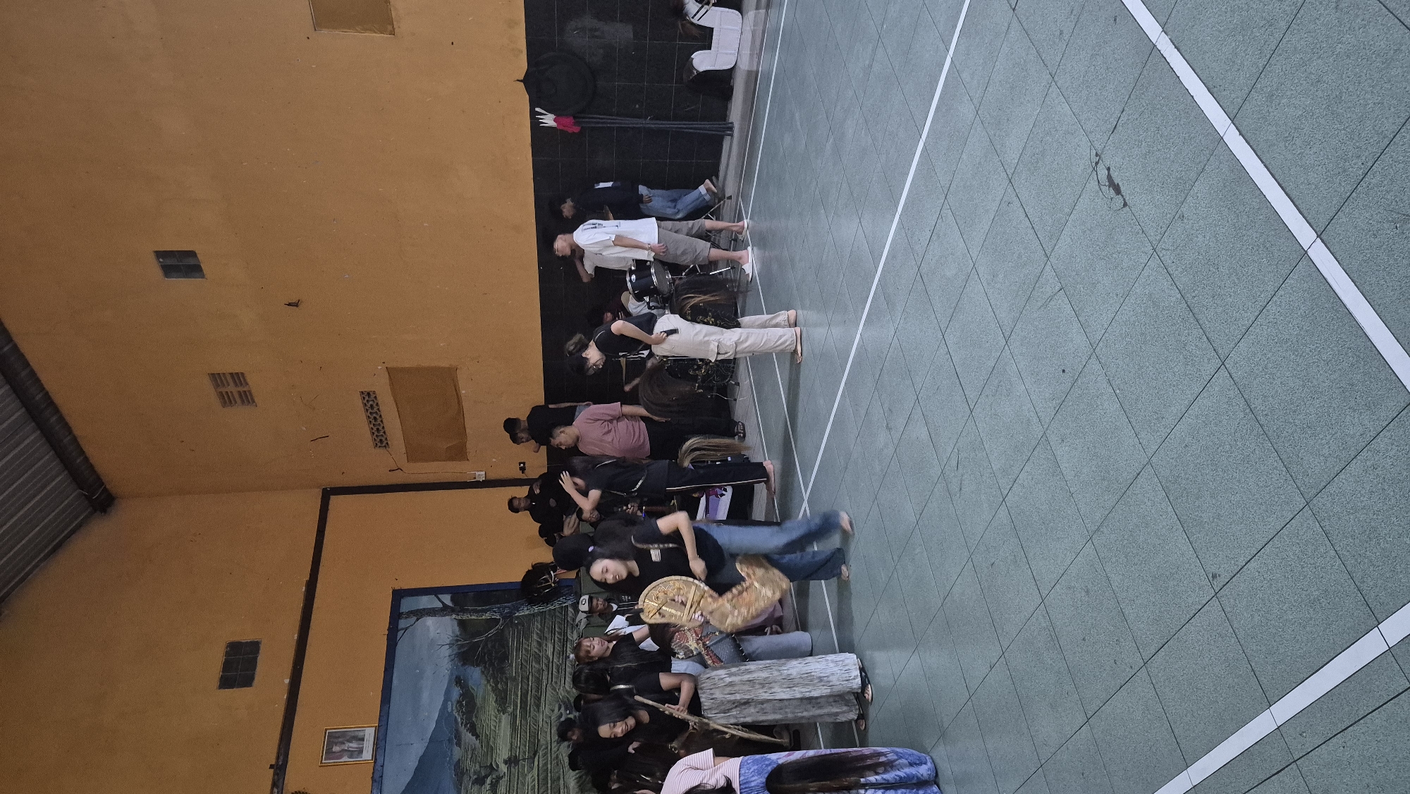 The image depicts an indoor gathering of people in what appears to be a community hall or sports facility in Sumowono, Indonesia. The hall has a corrugated metal roof and orange-painted walls. A large mural is visible on the left wall, depicting a landscape. Several individuals, mostly young adults, are present, some dressed in casual attire and others in traditional-looking outfits, including women wearing patterned skirts and holding large, drum-like instruments. There's a drum set set up in the background, suggesting a musical performance or rehearsal might be taking place. The floor is tiled in a green and white checkered pattern. Some individuals are interacting, talking and observing. The overall atmosphere seems casual and social.
