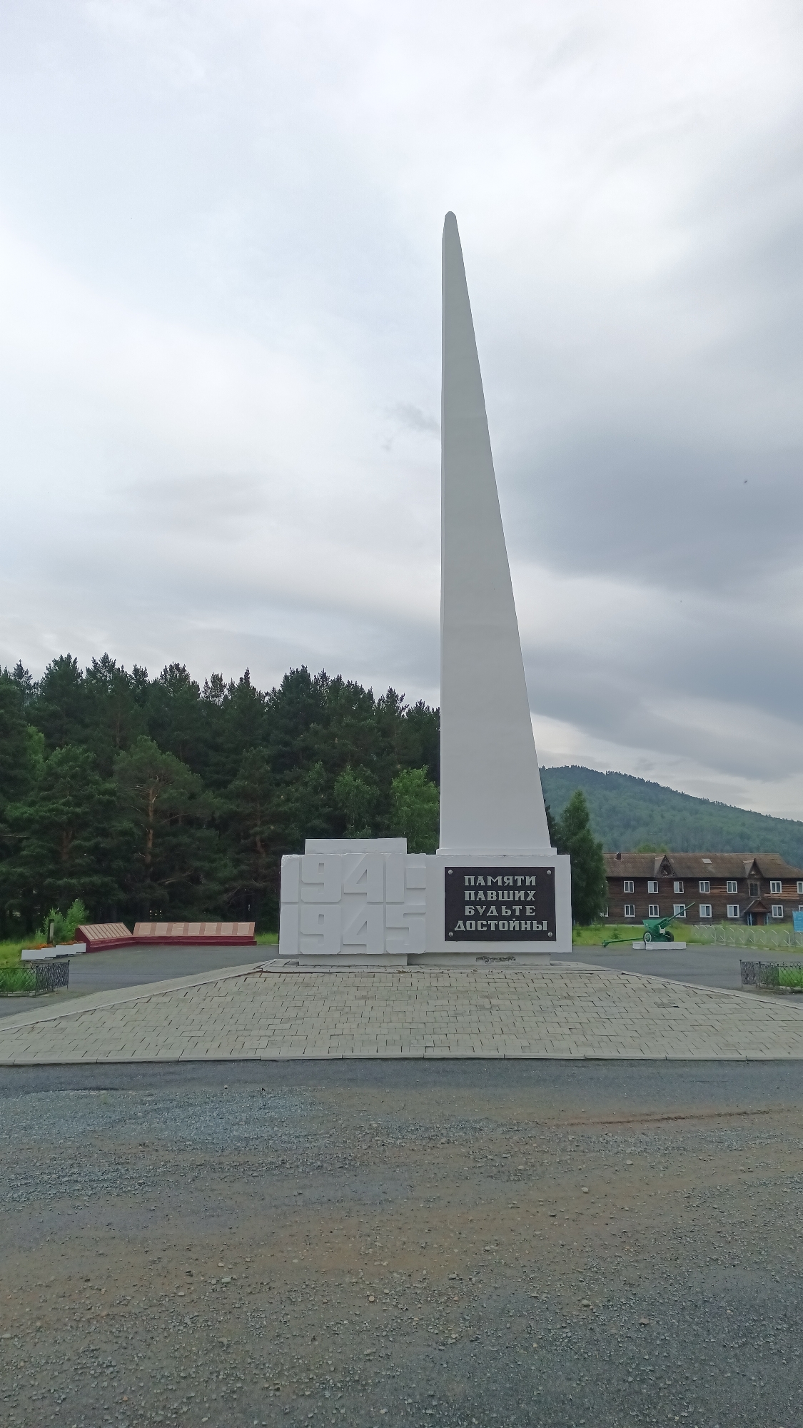 The image shows a white obelisk monument in the city of Abaza, Russia. The obelisk is tall and slender, tapering to a sharp point. At the base of the obelisk, there is a rectangular stone slab with the years "1941" and "1945" inscribed in large, raised block letters. Below this, a dark plaque contains Russian text that translates to "To the memory of the fallen, be worthy."

The monument is situated on a circular paved area, surrounded by a gravel path. In the background, a dense forest of green pine trees stretches across the landscape. Beyond the trees, several wooden buildings with pitched roofs and numerous windows are visible on a hillside. A small, green artillery piece is positioned to the right of the monument, near the buildings. The sky is overcast with gray clouds. There are no people visible in the image.