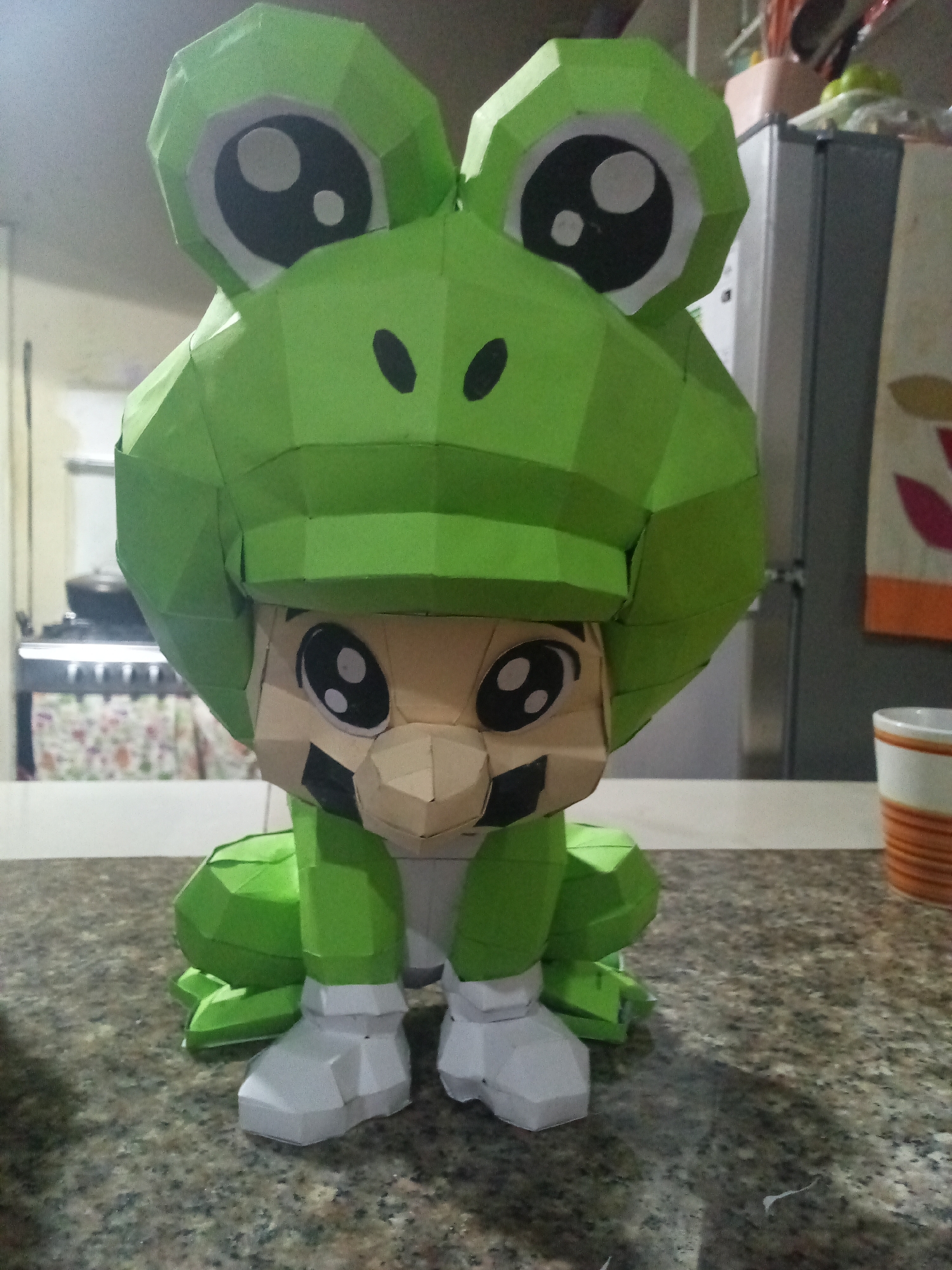 The media file displays a papercraft figure of Mario in his Frog Suit from the Super Mario Bros. 3 game, positioned centrally on a speckled dark grey and brown kitchen countertop. The figure is predominantly green, with the frog hood featuring large, cartoonish black and white eyes and two small black dots representing nostrils. Beneath the frog hood, a tan face with black mustache details and smaller black and white eyes is visible, suggesting the character Mario. The figure's body, arms, and legs are green, while the feet are white. The construction shows distinct polygonal facets characteristic of papercraft.
The scene is set indoors, likely in a kitchen in El Valle del Espíritu Santo, Venezuela. The background is slightly blurred, revealing various kitchen elements. To the left of the figure, a stove or oven with visible burners and a patterned tablecloth underneath can be seen. To the right, a silver-colored refrigerator with a patterned dishtowel hanging from its handle is present. A white mug with orange horizontal stripes sits on the countertop near the refrigerator. No specific activity is taking place, as it is a static image of the papercraft model. The lighting is consistent with an indoor environment. There is no visible text in the image.