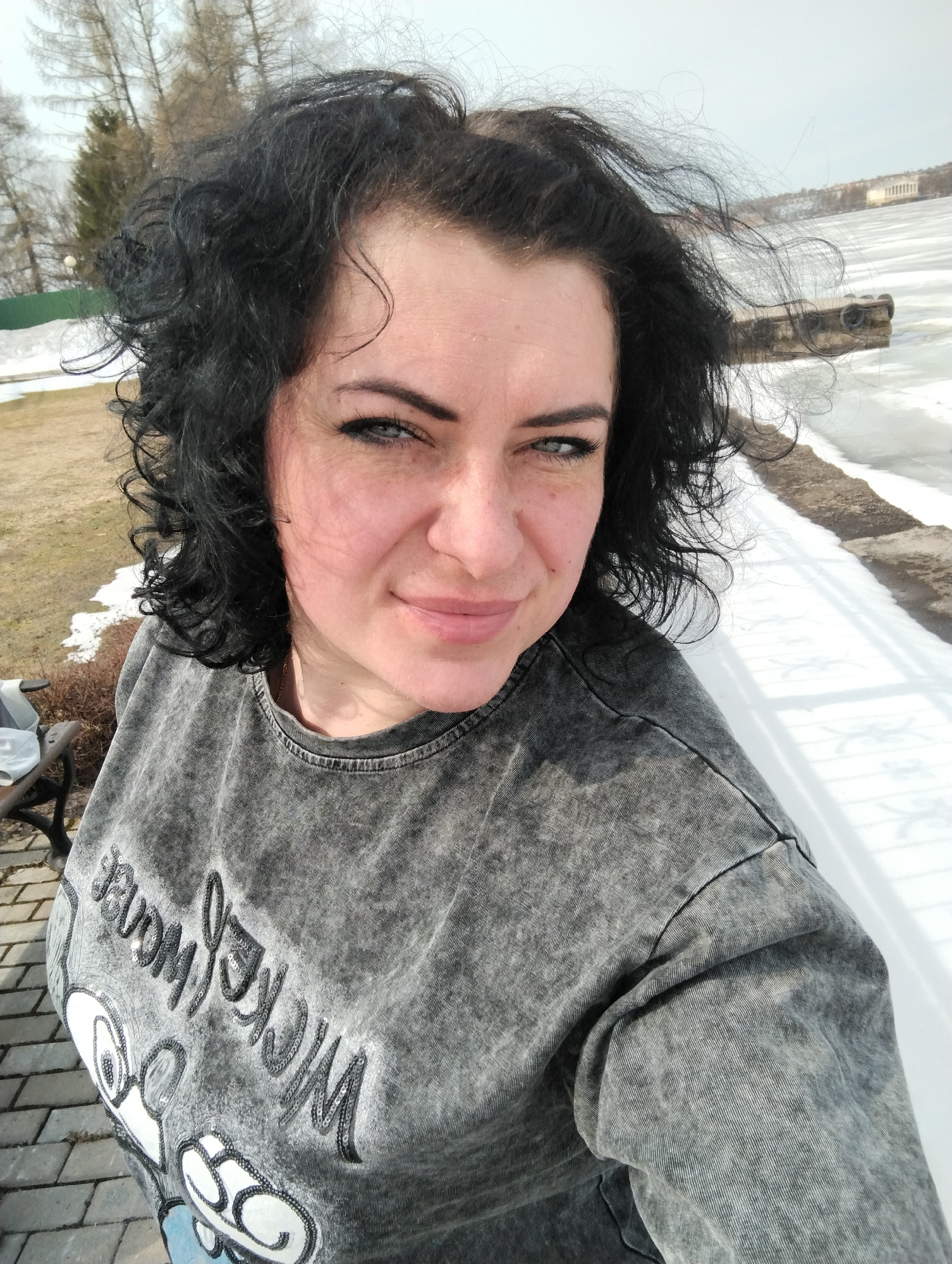 A woman with dark, curly hair is taking a selfie outdoors. She is wearing a grey sweatshirt with the words "Mickey Mouse" written on it, adorned with sequins. Her expression is a slight smile, and her eyes are light-colored.
The background shows a winter scene. There is snow and ice covering a body of water. A concrete barrier or ledge runs along the edge of the water. In the distance, a building can be seen on the opposite shore. To the left, there is a patch of grass with some bare trees. The time of day appears to be daytime due to the presence of sunlight.
The location is likely Votkinsk, Russia, given the prompt's direction. The body of water is most likely the Votkinsk Reservoir. The building in the distance could potentially be a landmark near the reservoir.