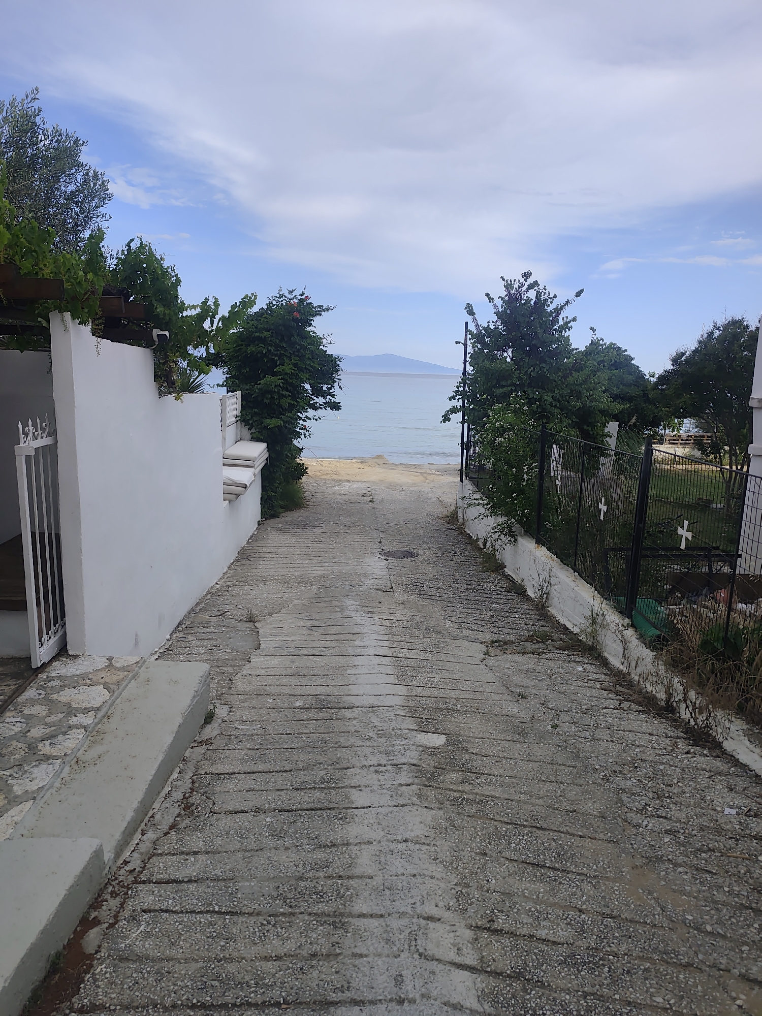 The image depicts a downward sloping concrete path leading towards a beach and the sea in Palio, Greece. On the left, a white wall borders the path, with a white gate and a small step leading into an unseen area. Lush green foliage and trees overhang the wall. On the right, a black metal fence encloses an area with green grass and more trees. The fence has decorative white crosses on it. The sea is calm and blue, with a distant landmass visible on the horizon. The sky is partly cloudy with a light blue hue. The path itself is weathered and appears to be made of textured concrete with visible expansion joints or grooves. There are no people visible in the image.