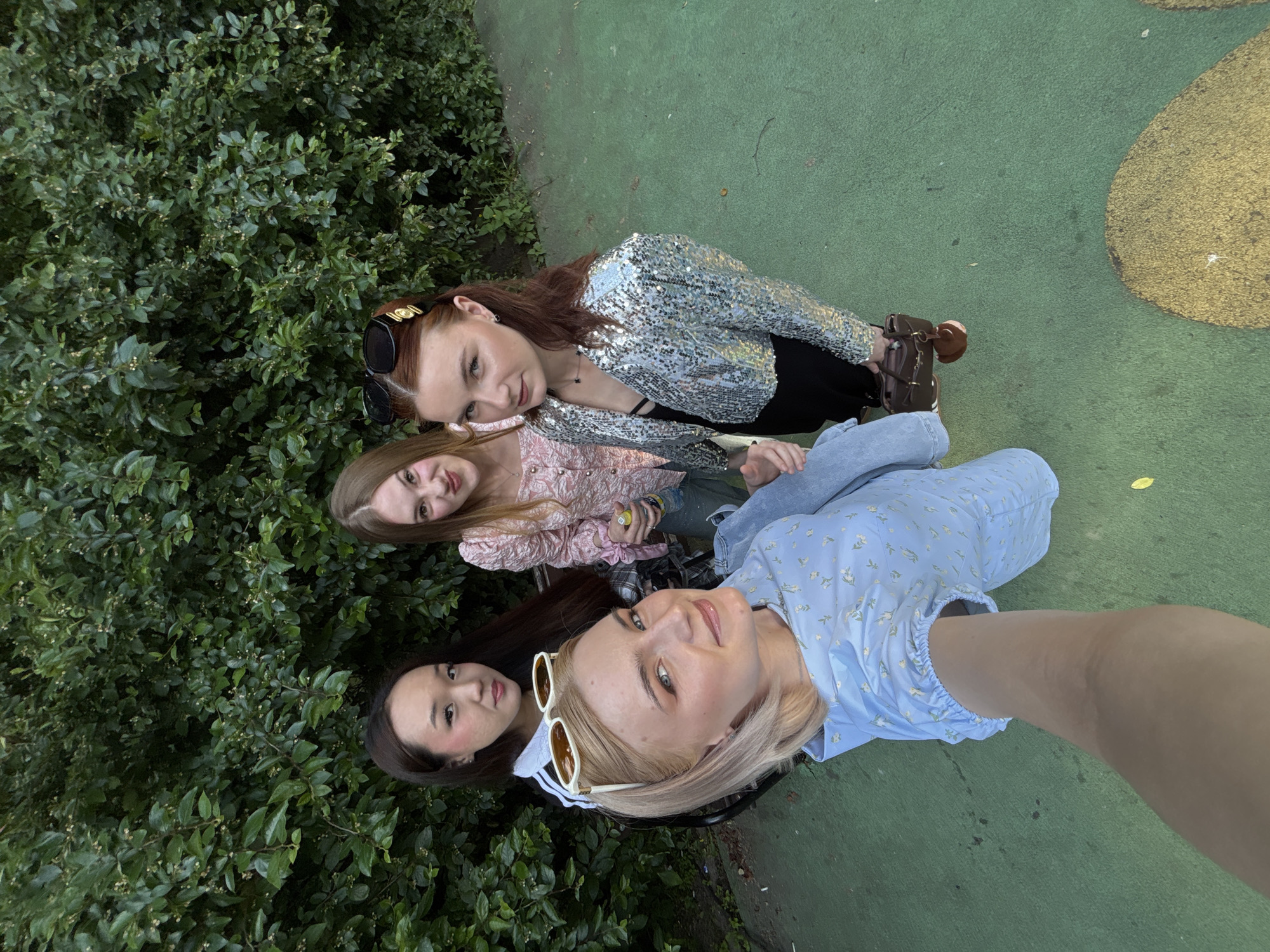 The image captures four young adult women posing outdoors in Vsevolozhsk, Russia. The woman in the immediate foreground, positioned slightly to the left, has blonde hair and is wearing a light blue dress with a subtle white floral pattern. She has white-framed sunglasses resting on top of her head and extends her right arm to hold the camera for a selfie, looking directly into the lens with a slight smile.
Behind her, to the left, a woman with dark hair and light skin is visible, wearing a black top with white stripes on the collar, consistent with an athletic brand design. She also looks towards the camera. To the right of the blonde woman, a woman with light brown hair is depicted, wearing a textured pink top. She holds a clear plastic bottle with a transparent liquid in her right hand and directs a slightly playful expression towards the camera.
The fourth woman, located to the far right, has reddish-brown hair styled with a gold-toned clip and black sunglasses on her head. She is wearing a cropped, long-sleeved jacket adorned with silver sequins over a black garment and black pants. She holds a dark brown leather handbag with a matching dark brown heart-shaped charm in her left hand, looking towards the camera.
The background consists of dense, dark green foliage, forming a continuous wall of vegetation. The ground beneath the subjects is a flat, green-colored paved surface, exhibiting some minor scuffs and marks. In the lower right quadrant of the image, a large, irregular yellow-gold shape is present on the green ground, indicating a painted or stained area. The scene is illuminated by natural light, producing soft, uniform illumination without harsh shadows.