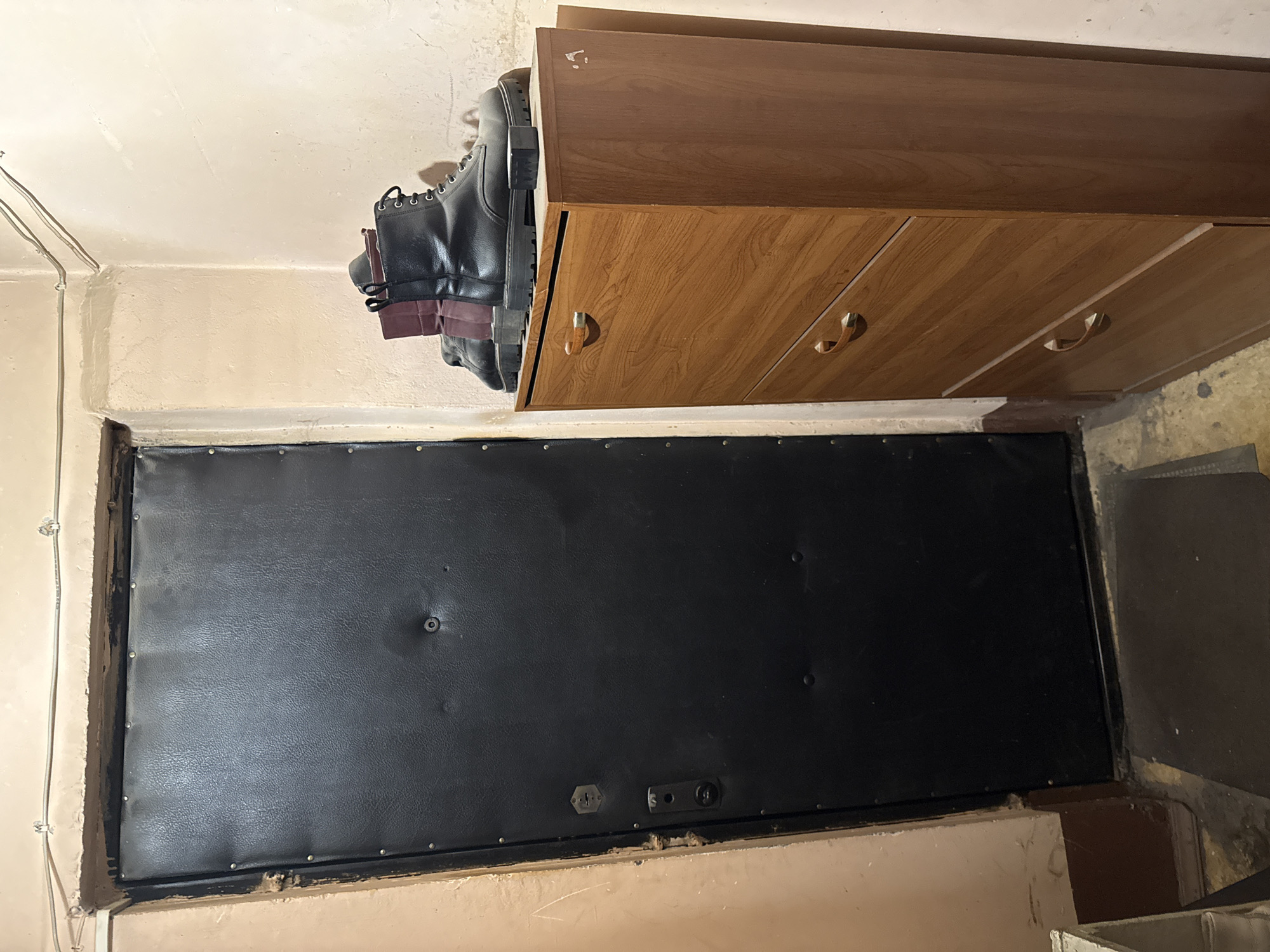 This image shows a close-up view of a dark, rectangular object, possibly a door or a panel, secured within a frame. The surface of the object is textured, with several visible indentations and what appear to be small metal studs or rivets around its perimeter. A circular latch or fastener is visible near the top center of the object, and below it, there are two other metal fixtures, one hexagonal and the other rectangular, that suggest a locking mechanism.
Above this dark object, a wooden cabinet with two drawers is mounted. The drawers have curved wooden handles. One of the boots, black with laces and a burgundy cuff, is placed on top of the cabinet, with its sole resting on the wood and its shaft extending upwards. The boots appear to be heavy-duty, possibly work boots or combat boots.
The surrounding environment is dimly lit, with a concrete floor visible in the lower right corner. The walls are painted a light, neutral color, with some visible imperfections. Wires are attached to the wall on the left side of the image. The overall impression is that of a storage area, utility space, or perhaps a basement. There is no clear indication of the time of day or weather conditions. The scene is static, with no activity taking place. No text is visible in the image.