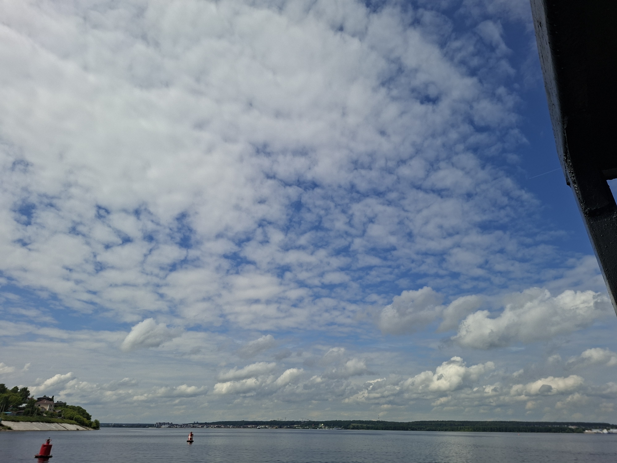 The image depicts a wide, calm body of water, likely a river or lake, under a bright blue sky dotted with numerous white, fluffy clouds. On the left bank, a verdant hill slopes down to the water, topped with several buildings, including what appears to be a large house or mansion surrounded by trees. A red buoy is visible in the foreground on the water, and another smaller, red and white buoy is further in the distance. Across the water, the opposite bank is lined with a distant cityscape and a dense line of green trees. In the upper right portion of the frame, a dark, angled structure, possibly part of a boat or bridge, partially obstructs the view. The overall scene suggests a sunny day with mild weather, likely in the city of Verkhny Uslon, Russia, given the context of the prompt.