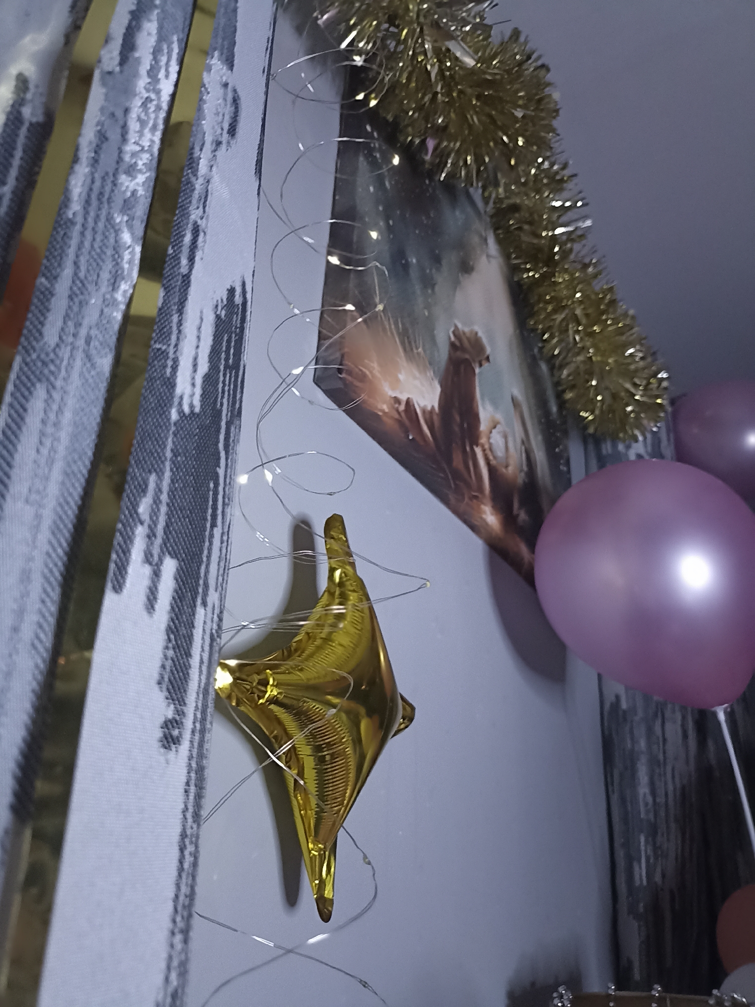 The image shows a close-up of a wall decorated with a gold star-shaped balloon, purple balloons, gold tinsel, and string lights. A framed picture is partially visible behind the tinsel. The scene is indoors, likely a home environment, with patterned fabric drapes or curtains on the left side of the frame. There is no indication of the image being located in Annagry, Ireland. The lighting suggests it is nighttime or a dimly lit interior.