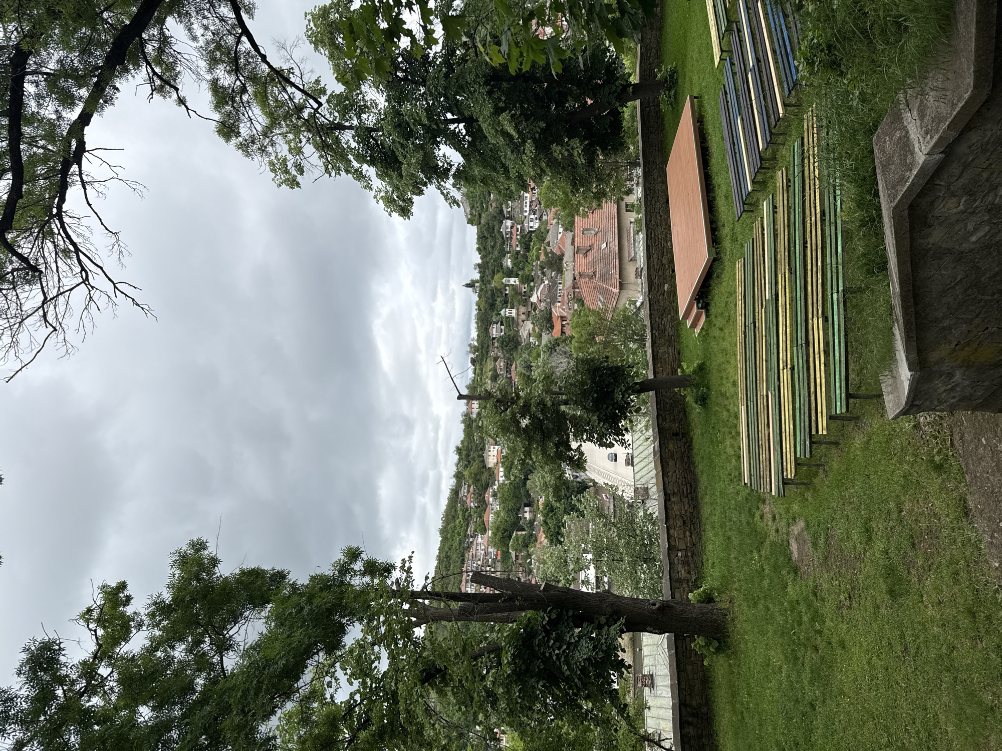 The image depicts an outdoor setting in the city of Lovech, Bulgaria. The scene is observed from an elevated perspective, looking down upon a landscape filled with houses and lush greenery. The sky is overcast. In the foreground, the ground is covered in grass. Adjacent to the grass, there are wooden benches that are painted in varying colors and arranged in rows, suggesting an outdoor theater or seating area. A wooden stage platform sits nearby. Stone walls are visible in the background, adding texture to the scene. Trees are strategically positioned, framing the image from the top and the sides. The trees are densely foliaged. The houses in the distance appear to be built on a hill. The overall composition suggests a scenic overlook of the city.