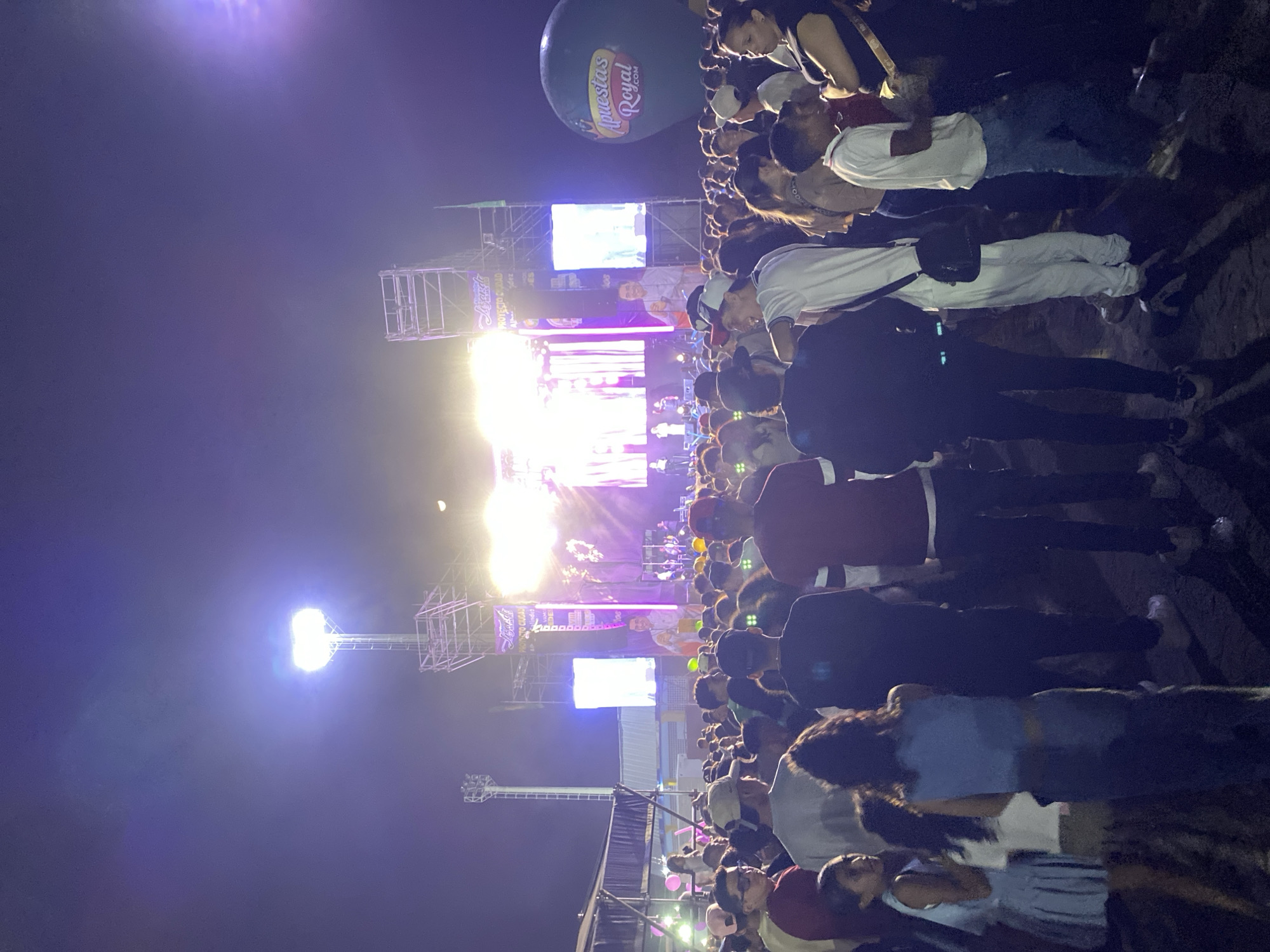 This image depicts a vibrant, crowded outdoor event at night in Tinaquillo, Venezuela, likely a concert or public festival. A large crowd of people, mostly adults dressed in casual attire, are gathered, facing a brightly lit stage.
The stage features a prominent metal truss structure, adorned with numerous spotlights and large LED screens. These screens display various content, including a man's face and what appears to be branding or text. One partially visible banner on the stage structure reads "Semana Royal" or similar, with "Royal" being clearly visible. The area is heavily illuminated by stage lights and tall floodlights, casting bright glows against the dark night sky. A crescent moon is also visible high above the scene.
Floating above the crowd is a large, dark green inflatable balloon or blimp, prominently displaying the text "Apuestas Royal .com", indicating a key sponsor or event organizer. The overall atmosphere suggests a lively and well-attended public gathering, with attendees focused on the performance or activity taking place on the stage.