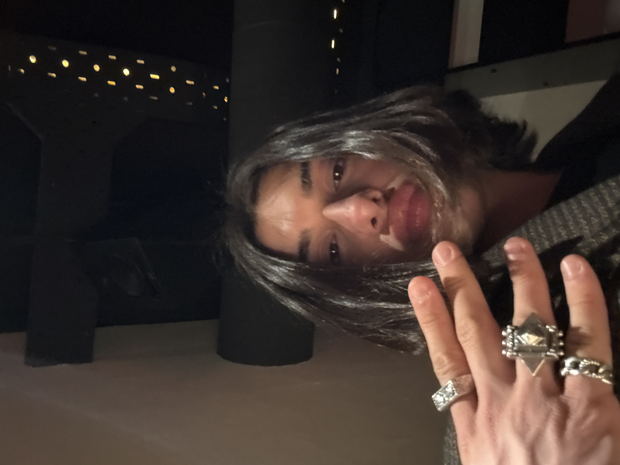 A person with dark hair and pale skin is visible from a close, canted angle, their face tilted towards the camera. They are sticking out their tongue. Three rings are visible on their left hand. The background features a dark, modern interior with blurred lights suggesting an urban setting. The location is unknown, there is not enough information to determine if it is in Yerevan, Armenia.