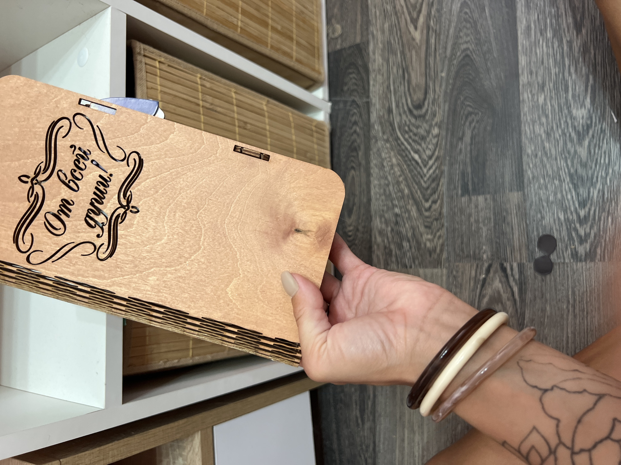 A right hand is holding a rectangular, light-brown, wooden box with Cyrillic writing and decorative engraving on the top left corner. The hand has pale skin, with visible veins, and three bangles on the wrist in cream, light-brown, and dark-brown. A tattoo is visible on the forearm. The floor has a wood-grain texture. In the background is a white shelf unit with tan wicker drawers. The location is inferred to be Boguchar, Russia, based on the Cyrillic writing. Two small black circles are also visible.