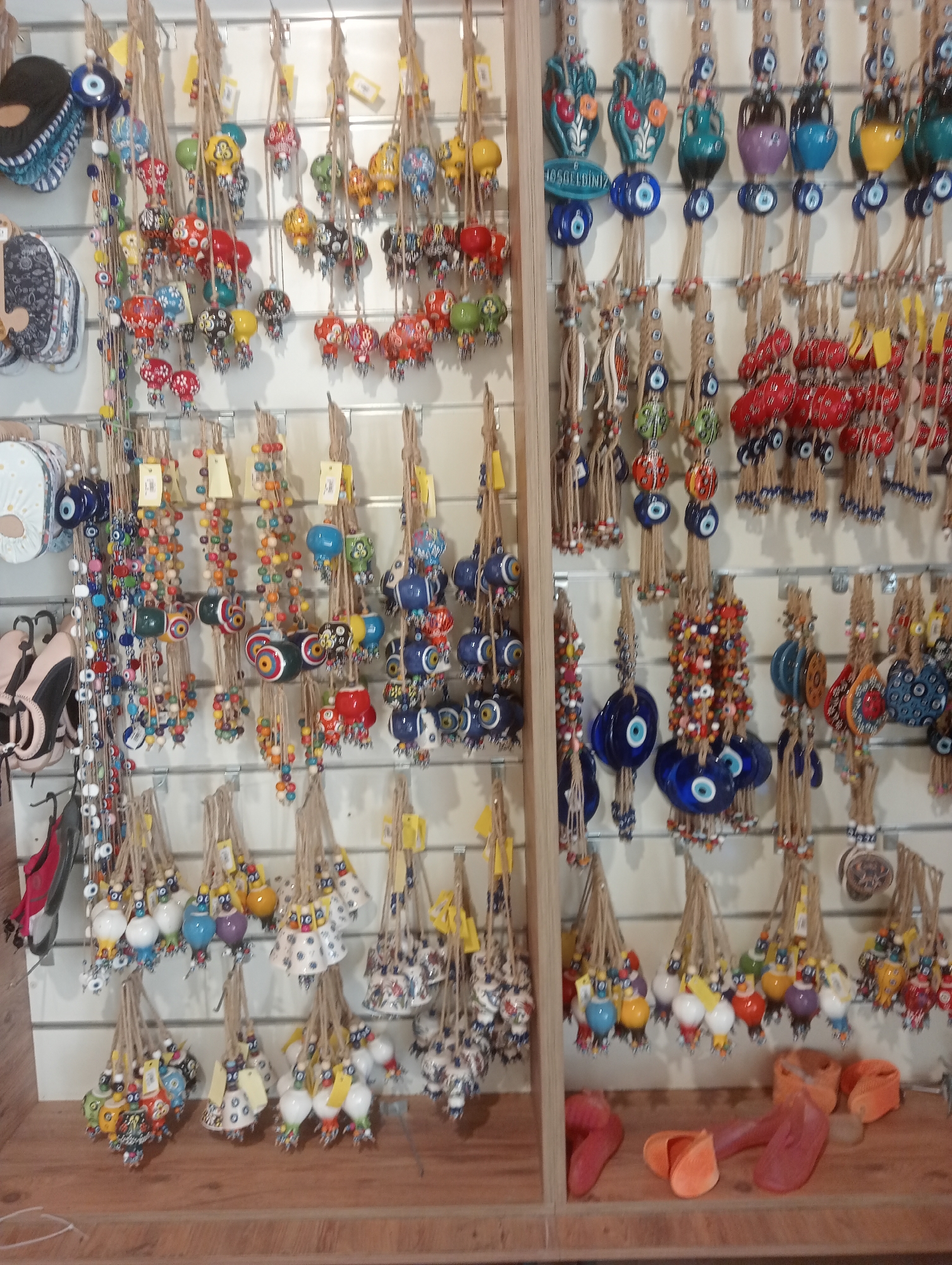 The image shows a display of colorful hanging ornaments, possibly souvenirs, in a shop. The ornaments are diverse, featuring glass evil eye charms, ceramic figurines shaped like birds and owls, and decorative vases. They are suspended by rope or string against a white wall. Some of the ornaments have intricate patterns and vibrant colors. The display is well-lit, and the items are arranged in rows and clusters. In the foreground, on a wooden shelf, are several pairs of flip-flops in shades of pink and orange. The overall scene suggests a retail environment selling decorative items. The specific location is a shop in the city of Korkuteli, Turkey, known for its traditional crafts and local products.