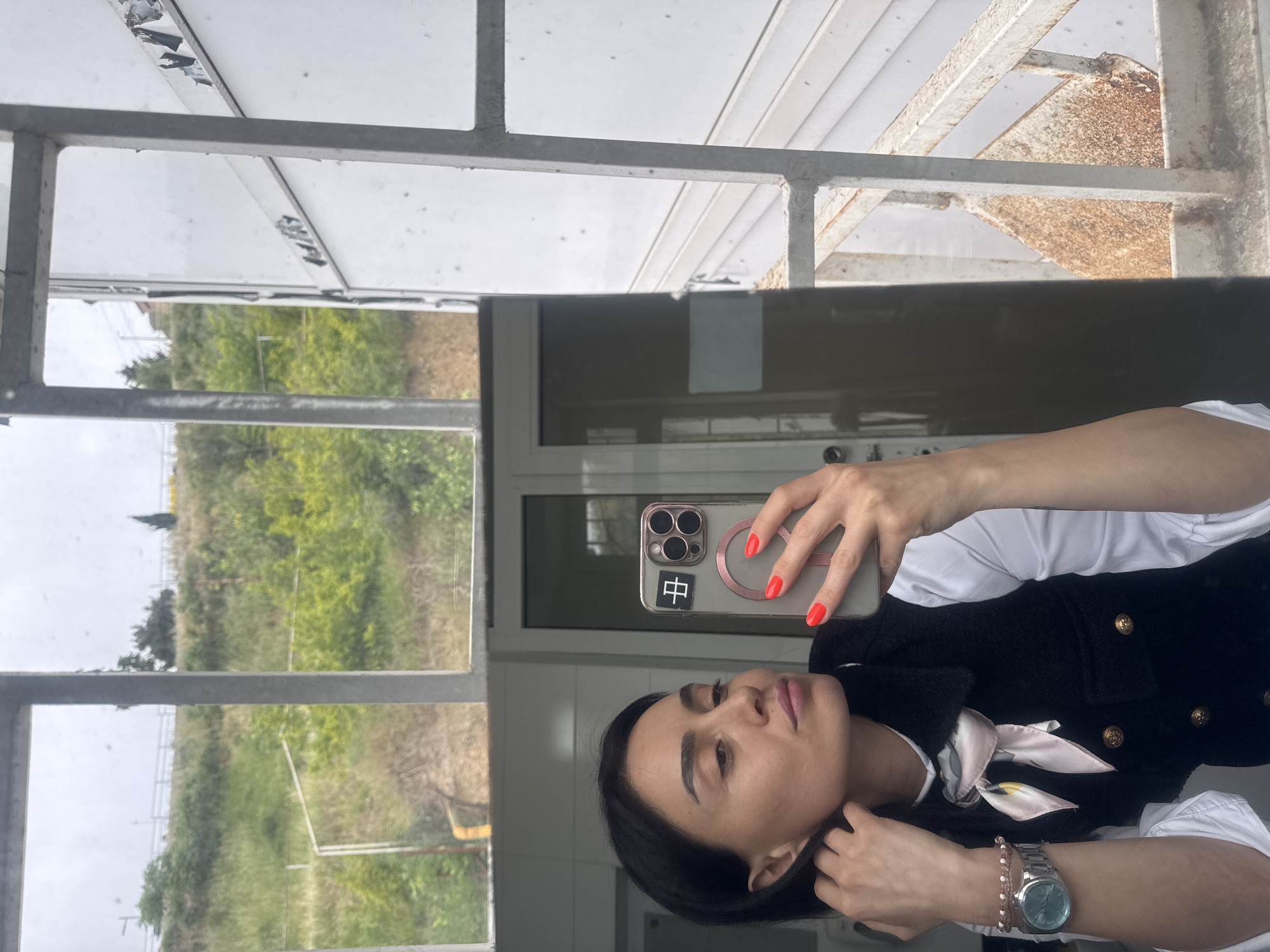 The original image is oriented 90 degrees clockwise, depicting a woman taking a selfie in a reflective surface, likely glass or a mirror. The woman, whose reflection is captured from the chest up, has dark hair and a neutral to slightly contemplative expression as she looks upwards. She is dressed in a white collared shirt, a black textured vest adorned with gold buttons, and a pink and white patterned scarf tied around her neck. Her left hand holds a silver-cased iPhone, which features a prominent triple-lens camera module and a black sticker displaying the Chinese character "中". Her fingernails are painted a vibrant red. On her right wrist, she wears a silver watch with a light-colored face and a beaded bracelet.
The background, visible through and beyond the reflective surface, reveals an outdoor scene under an overcast sky, suggesting daytime. A grey metal framework, possibly part of a large window or a canopy structure, shows signs of weathering and rust in places. Beyond this structure lies a landscape of green and brown vegetation, indicating a natural or semi-wild area. The setting appears to be a semi-enclosed outdoor space, such as a balcony or a viewing platform. While the location context is Tbilisi, Georgia, the image itself does not contain specific visual identifiers for this city.