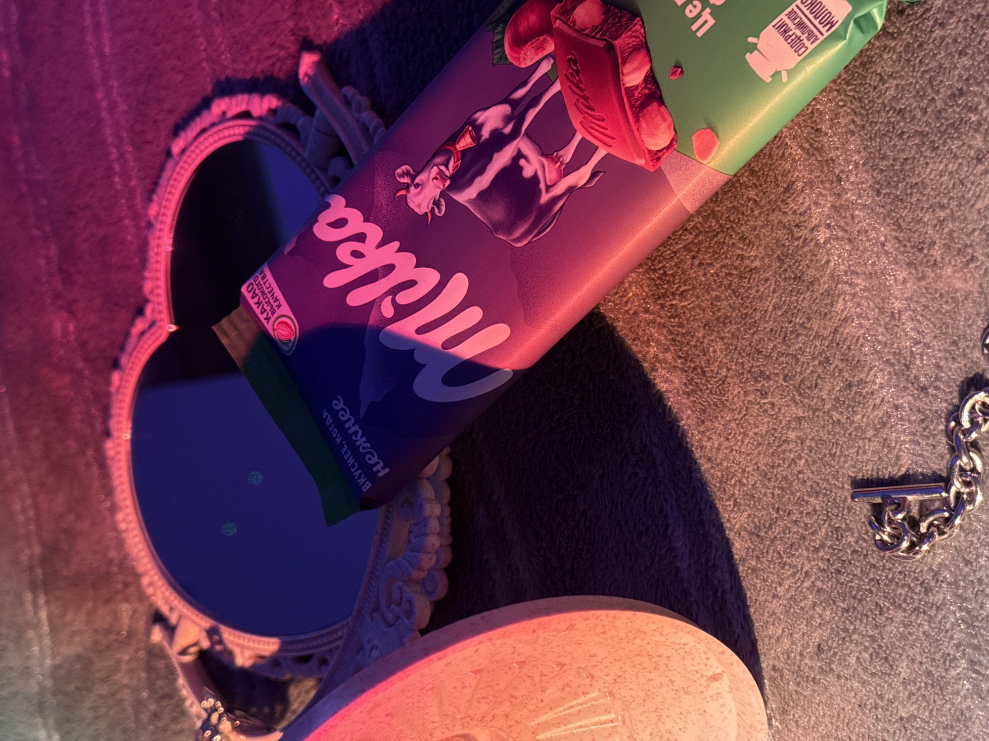 Here's a detailed description of the image:
**Scene:** A still life arrangement with a variety of objects under unique lighting conditions, likely indoors.
**Objects:**
* **Milka Chocolate Bar:** A purple and green Milka chocolate bar is prominently placed, angled diagonally.
* **Mirror:** An ornate, heart-shaped mirror with a vintage-style frame is positioned on the left side of the frame, reflecting a dark or obscured image.
* **Lamp:** The bottom of a lamp with the light on is placed on the bottom of the frame.
* **Chain Bracelet:** A chunky silver chain bracelet with a T-bar closure is lying on a surface with a visible fabric texture.
**Environment:**
* **Lighting:** The scene is lit with a mix of red and purple hues, casting shadows and creating a dramatic effect.
* **Surface:** The objects are placed on a textured fabric surface, appearing plush or carpet-like.
**General Notes:** The lighting and arrangement of objects give the scene an artistic, almost staged quality.
**Location:** Unknown, Unknown.