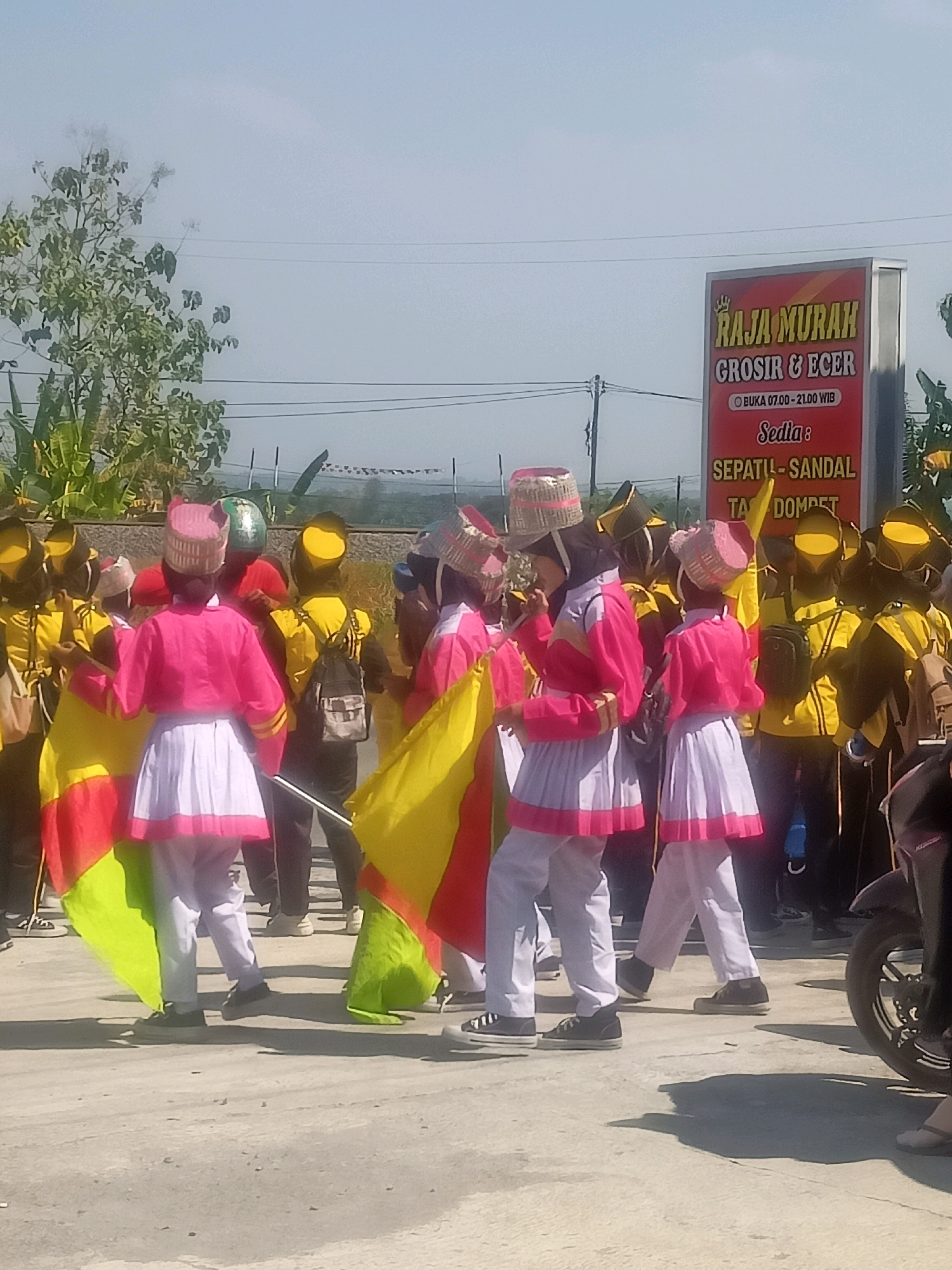 In the city of Sumberlawang, Indonesia, a group of individuals, predominantly young people dressed in bright uniforms, are participating in what appears to be a parade or performance. The attire of the participants includes distinctive pink and yellow jackets, white skirts, and elaborate hats. Some are holding flags in vibrant colors like yellow, red, and green, suggesting a festive occasion.
The background reveals a clear, hazy sky and a few trees, indicating an outdoor setting during daylight hours. A prominent red sign in the background displays text in Indonesian, advertising "RAJA MURAH GROSIR & ECER" (Cheap King Wholesale & Retail) and listing opening hours and available items like shoes, sandals, and wallets. The ground appears to be a paved surface, possibly a road or plaza. The presence of a motorcycle parked on the right side of the frame suggests it is a public area where vehicles are common. The overall impression is one of a lively community event, possibly a cultural or promotional activity.