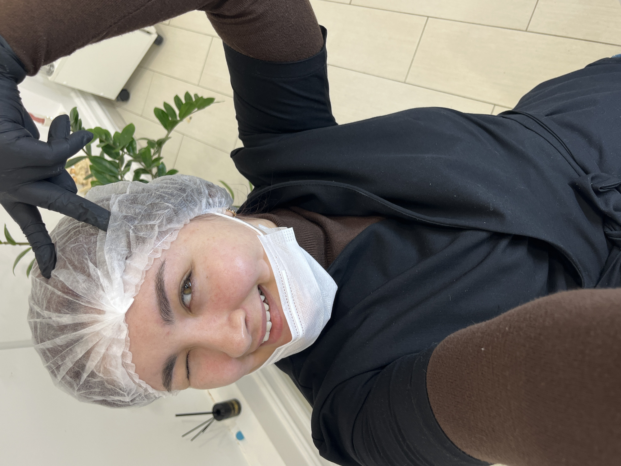 The scene depicts an individual inside an indoor professional setting, specified as being in Neftekamsk, Russia. A female subject, with fair skin and brown eyes, is centered in a close-up selfie-style shot. She is winking with her left eye and smiling broadly, revealing her upper teeth. Her head is covered by a white disposable hairnet, and a white disposable surgical face mask is pulled down beneath her chin, exposing her mouth and nose. She wears a dark brown long-sleeved undershirt beneath a black, wrap-style uniform top. Her left hand, adorned with a black disposable glove, is raised with the index and middle fingers extended in a "V" gesture near the top of her head. The background on the left features a light-colored wall with a small black diffuser containing reeds on a white ledge. To the right, a green leafy plant is partially visible, along with a white mobile cabinet on wheels. The floor consists of light-colored square tiles.