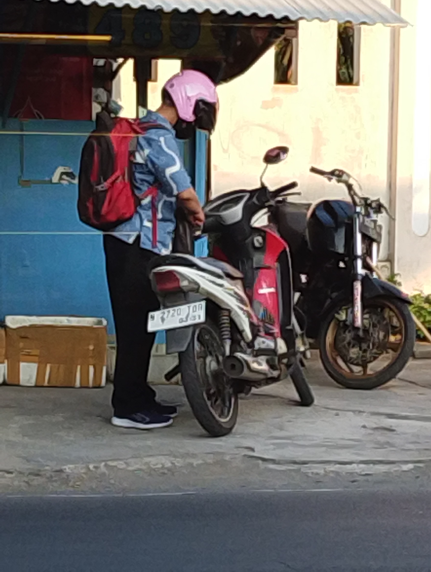 A person wearing a pink helmet, a blue patterned shirt, black pants, and dark blue athletic shoes stands next to two motorcycles on a paved surface. The person has a red backpack with black straps. One motorcycle is white with red and black accents, and the other is black with red accents. The white motorcycle has a license plate with the characters "N 2720 TON". In the background, there is a blue wall, a corrugated metal roof with yellow markings, and a beige cardboard box with tape. The scene appears to be outdoors, possibly on a street or sidewalk in Manaruwi, Indonesia.