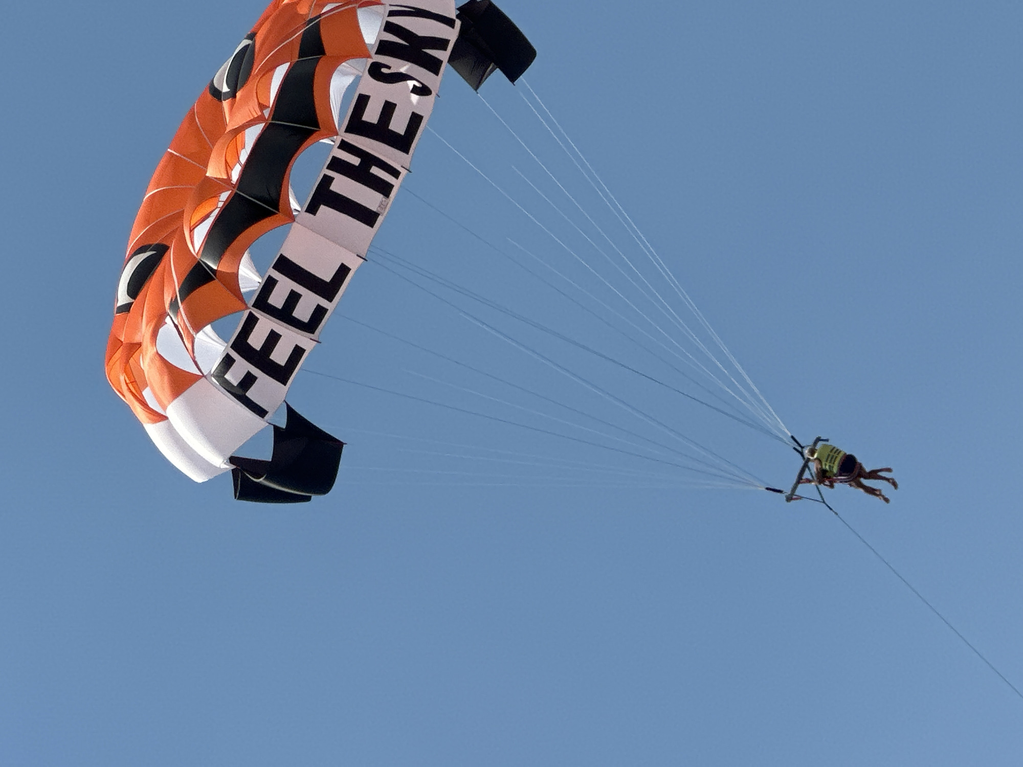 The image shows two people parasailing in a clear blue sky, with a large orange, black, and white parachute overhead. The parachute has the words "FEEL THE SKY" printed in large black letters. The two people are harnessed together, with one person appearing to be an instructor and the other a passenger. They are both wearing life vests. The scene is set in a bright, sunny day, likely during daylight hours. The location is identified as Boreti, Montenegro. The parasailing activity implies a coastal or lake setting, though water is not visible in the frame. The overall mood is one of adventure and freedom as they glide through the air.