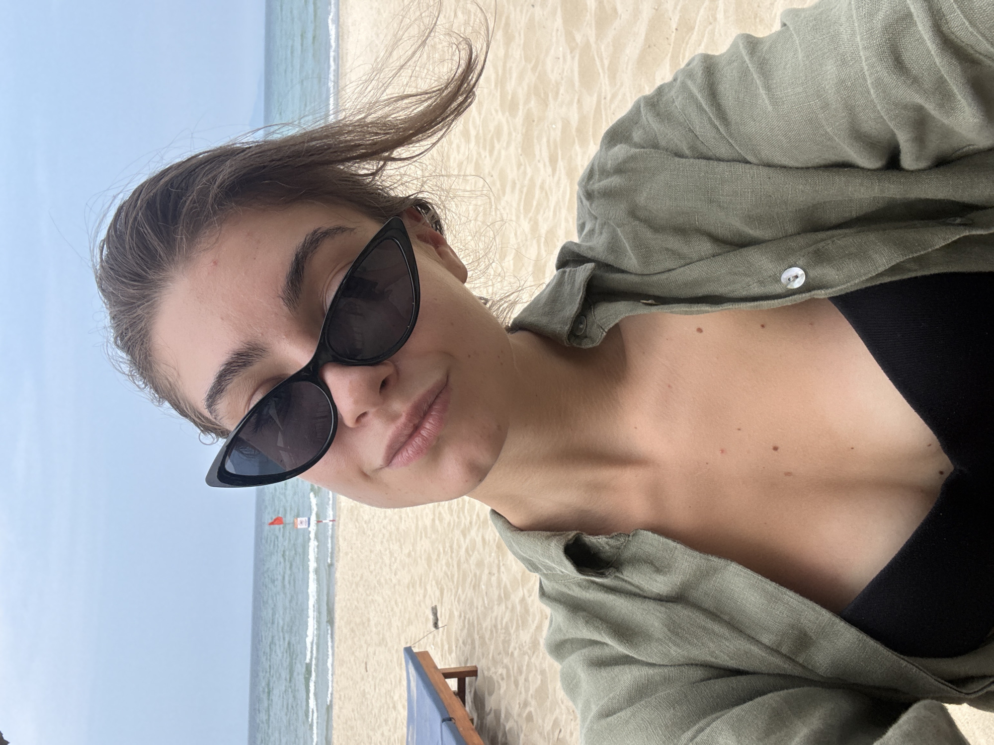 A young woman takes a selfie at a beach in Đà Nẵng, Vietnam. The image is oriented diagonally, with her face occupying the upper left quadrant. She is wearing large black cat-eye sunglasses that obscure her eyes, a loose-fitting, unbuttoned olive green linen shirt, and a black top or swimsuit underneath. Her brown hair is slightly tousled, suggesting a light breeze. She looks directly at the camera with a relaxed expression, featuring a subtle closed-mouth smile.
The background shows a sandy beach transitioning into the ocean with gentle waves, under a bright, clear sky, indicating daytime. A partial view of a blue and wooden beach chair is visible on the sand behind her. Further out in the water, a red flag on a pole can be seen. Her décolletage and neck are visible, showing several small moles. The image conveys a serene and relaxed atmosphere characteristic of a beach vacation. No discernible text is visible.