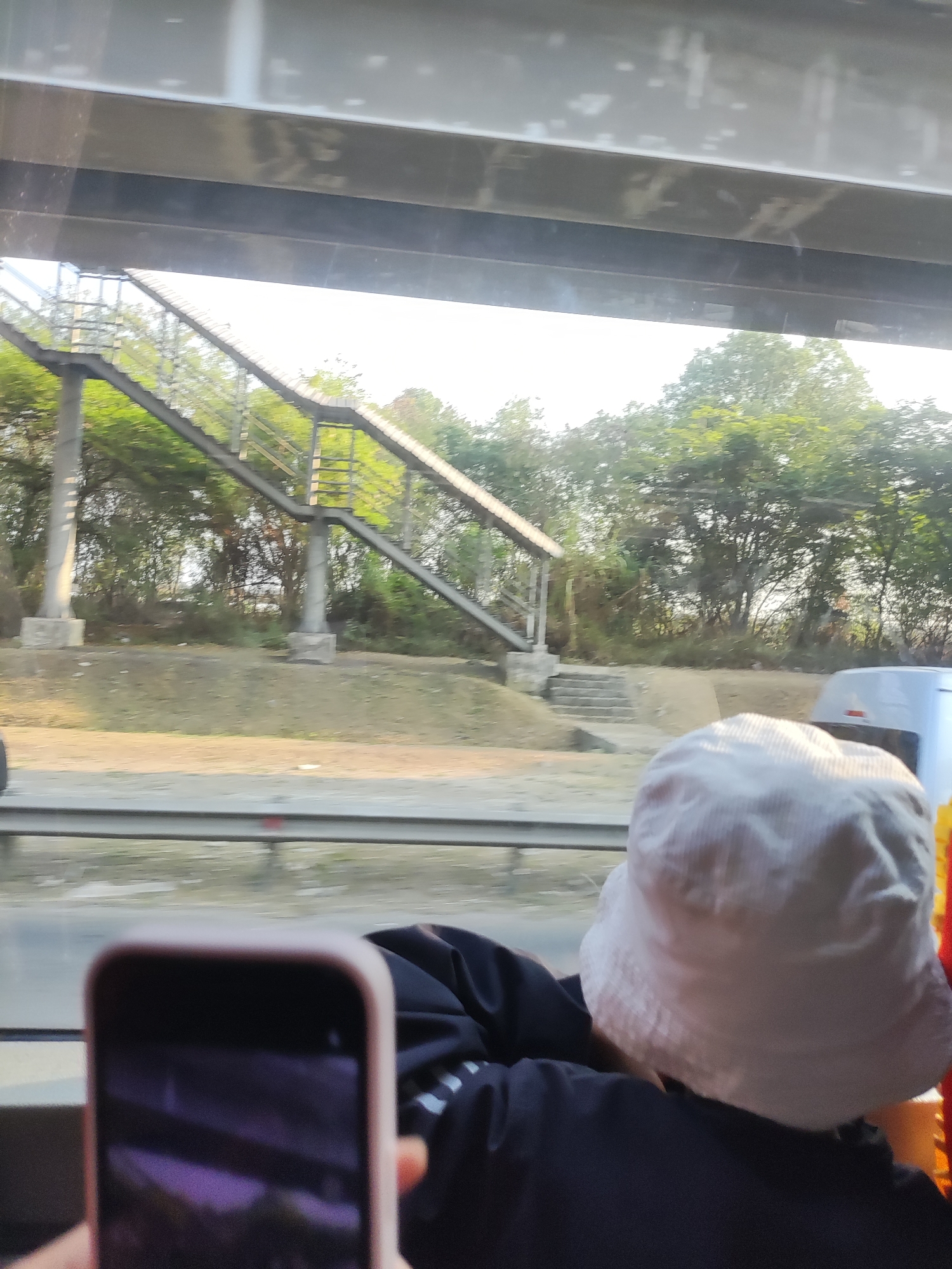 The image captures a view from inside a vehicle, looking out through a window. In the foreground, a person wearing a white bucket hat is visible from the back, with their shoulders and the back of their head in frame. They are wearing a dark jacket with white striped accents. To the left of the person, a hand holds up a smartphone, displaying a purple-tinted screen reflection.
Beyond the person and the vehicle's window, the scene opens up to an outdoor environment. A pedestrian overpass with metal stairs and railings is prominent, supported by concrete pillars. The overpass crosses over a road or pathway, bordered by a metal railing. Green trees and foliage form a dense backdrop behind the overpass, suggesting a location with abundant vegetation. The lighting indicates it is daytime, possibly late afternoon, with sunlight casting a warm glow on the scene. The overall impression is a transient view of an urban or semi-urban landscape, possibly along a road in the vicinity of Jayamukti, Indonesia, given the context. The image appears to have been taken while in motion, as indicated by slight motion blur in the background elements.