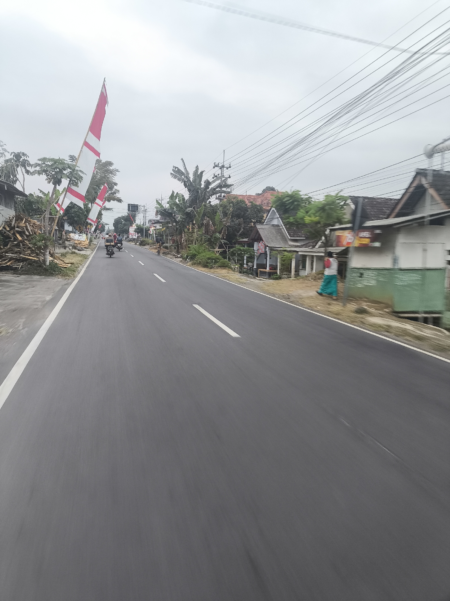 The image captures a view from a moving vehicle on a paved road in Purworejo, Indonesia, under an overcast sky. The road is divided by a white line. On the left side of the road, a pile of cut wood is visible next to a building. Several Indonesian flags, with red and white horizontal stripes, are mounted on poles along the roadside. In the distance, a few motorcycles with riders are seen on the road. To the right of the road, houses with tiled roofs are present. One house has a green-painted section, and a person wearing a teal-colored sarong is standing near it. Overhead, a complex network of electrical wires stretches across the sky.