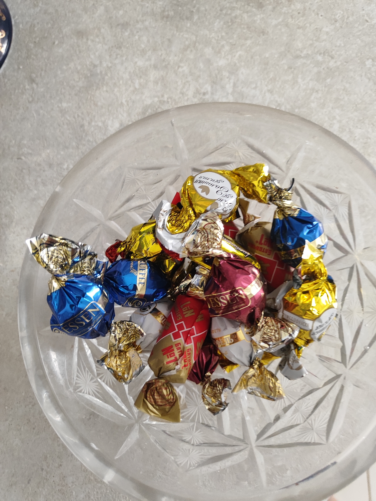 A pile of individually wrapped chocolates sits in a clear, intricately patterned glass bowl. The chocolates are wrapped in various colored foils, including gold, blue, and burgundy. Some wrappers have visible text and logos. The bowl is placed on a light-colored, textured surface. The lighting suggests an indoor environment. The scene is shot from a top-down perspective.