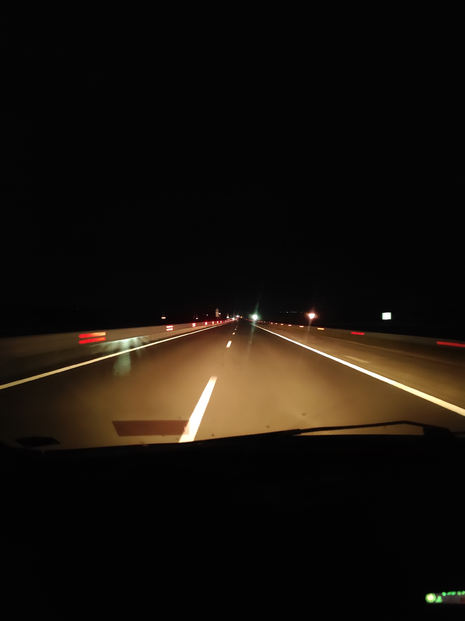 The image depicts a view from inside a vehicle traveling on a highway at night. The road is illuminated by the vehicle's headlights, casting a warm glow on the wet asphalt. White lines mark the lanes, and red reflectors are visible along the shoulder. Distant lights suggest other vehicles and possibly signs in the darkness. The sky is completely black, indicating a moonless night or heavy cloud cover. The foreground shows the car's dashboard, with a windshield wiper partially visible. There are no people or other subjects in the frame. The overall scene is one of solitary travel through a dark, unknown landscape.