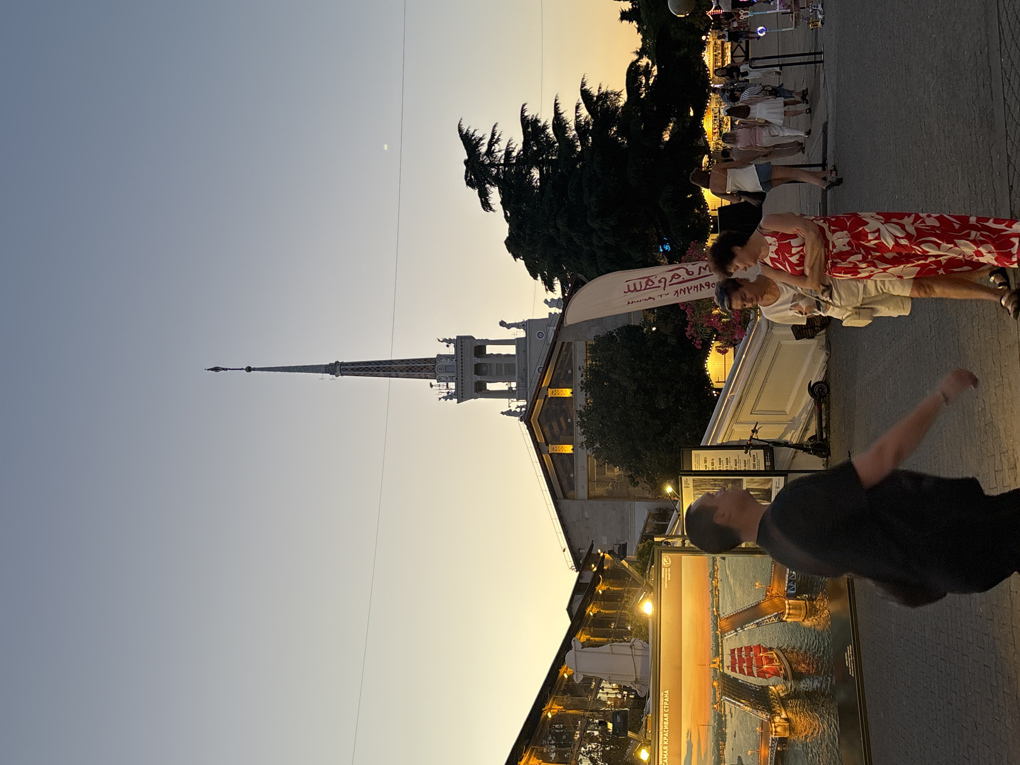 The image depicts a street scene in Sochi, Russia, during sunset. In the background, a tall, spire-like building with decorative elements and statues stands prominently against the pale sky. To the left of the spire, a long, multi-story building with large windows illuminated by warm lights lines the street. In the foreground, a man in a black t-shirt walks away from the camera. To the right of the man, two women are standing and talking. One woman wears a white t-shirt and shorts, while the other is in a long red and white floral dress. Further in the background, several people are walking, and market stalls with lights are visible. A large framed artwork featuring a cityscape with a red-sailed ship is displayed on the left side of the image. An electric scooter is parked near a white railing. The overall atmosphere suggests a lively evening in a city square.