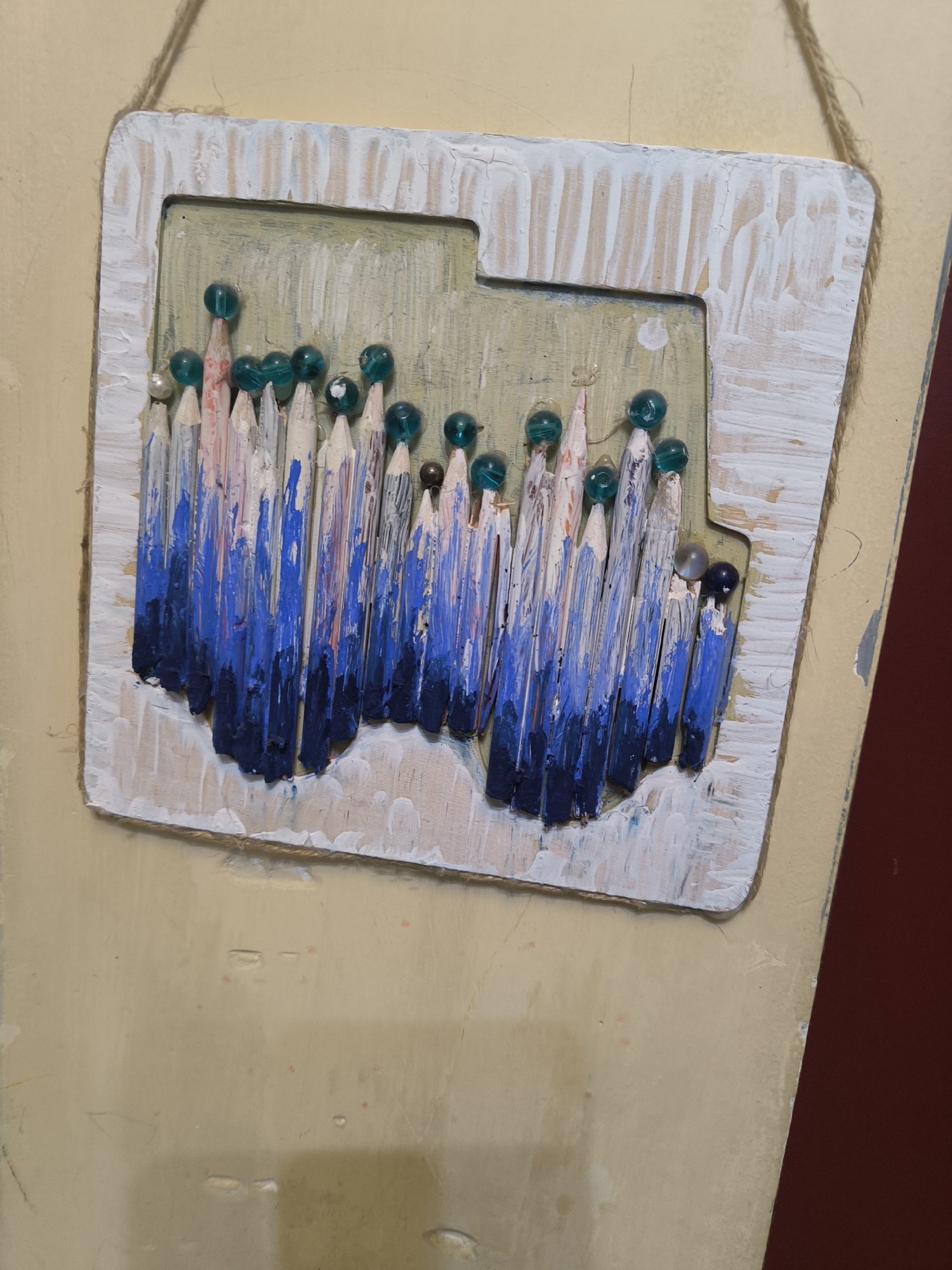A handmade art piece is hanging on a light yellow wall, possibly in Tbilisi, Georgia. The artwork features vertically arranged, painted, partially colored pencils of varying lengths with their tips facing down. The lower portions of the pencils are covered in dark blue paint, which fades into white at the top. The top of each pencil is topped with a small, spherical, glass-like bead, mostly blue-green in color, with one being white and another being brown.
The pencils are mounted on a light-colored wooden board with a distressed white-painted frame. The board is beige in the center area. A thin rope is attached to the top corners of the frame, creating a hanging mechanism. The background wall is a muted yellow, and a portion of a dark red or brown surface is visible to the right of the artwork.