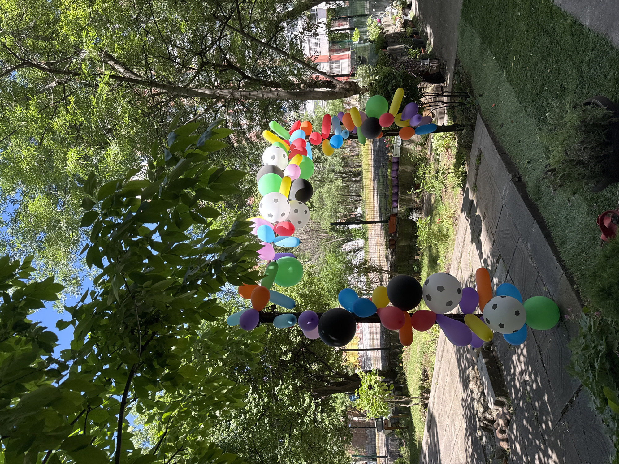The scene depicts a colorful balloon archway erected in what appears to be a garden or park setting. The archway consists of an assortment of balloons of various colors and shapes, including soccer ball-patterned balloons and elongated balloons. The archway is positioned above a pathway paved with rectangular stones. The pathway is flanked by grassy areas and foliage, suggesting a well-maintained outdoor space.
In the background, a residential or commercial building with multiple stories can be observed. Based on the architecture and the presence of greenery, it is plausible that this photograph was taken in a park within a city, such as Sofia, Bulgaria, given the architecture style. However, without further information such as local landmarks or signage, confirming the exact location in Sofia is impossible. The image includes trees and a partially visible sky. No individuals are clearly visible in the image.