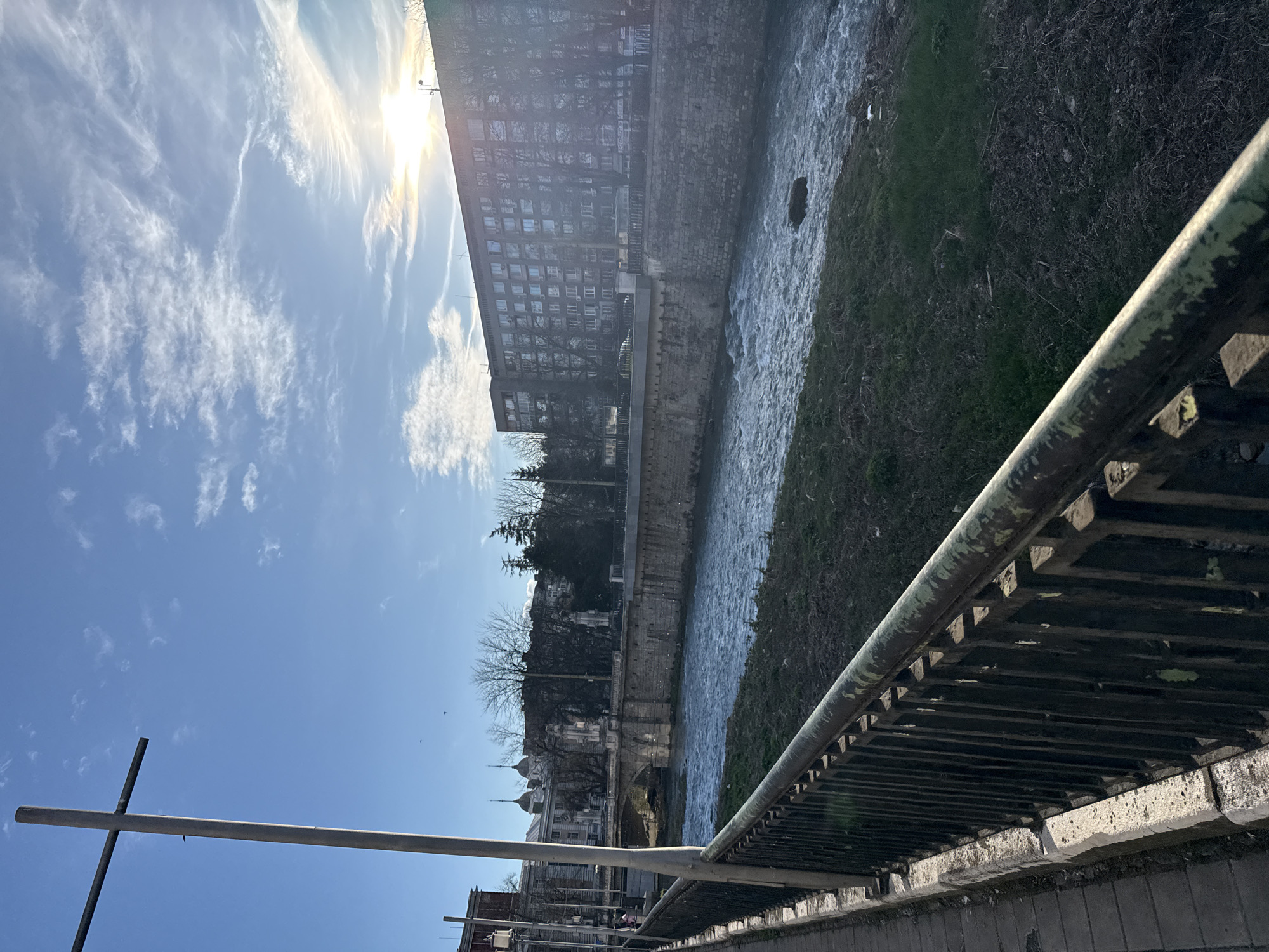 The image depicts a river scene in Gabrovo, Bulgaria, taken from a bridge. A river flows between concrete and stone embankments. On the left bank, a large, multi-story building dominates the view, casting a shadow. Further downstream, traditional Bulgarian architecture with domes and spires is visible. On the right bank, grass and vegetation grow along the river's edge. A metal railing runs along the bridge, partially obscuring the view. The sky is blue with scattered clouds and the sun shining brightly. The photograph is taken from an elevated perspective.