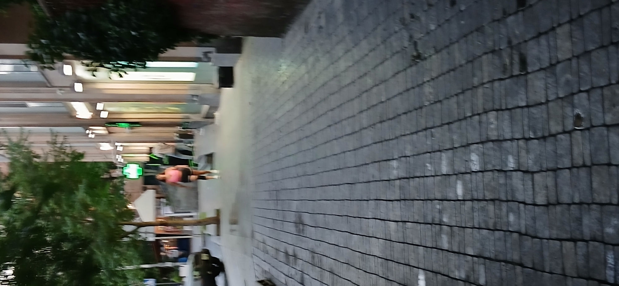 In Yerevan, Armenia, on a sidewalk paved with gray rectangular stones, a person is captured mid-stride, walking away from the camera. The individual, wearing a bright pink top and dark shorts with knee pads, is heading towards a building facade lined with illuminated windows and storefronts. A green cross sign is visible on the left side of the building, indicating a pharmacy. Trees with green foliage line the left side of the street, and a dark-colored vehicle is parked on the far left. The scene appears to be in an urban setting, with the perspective emphasizing the paving stones and the receding figure.