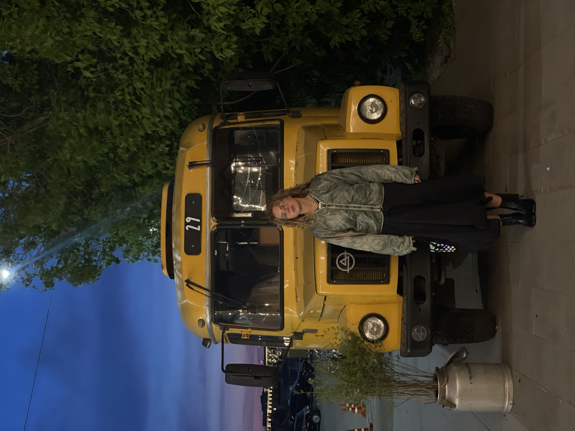 The image depicts a fair-skinned woman standing in front of a vintage yellow bus. She is wearing a light-colored patterned jacket, a dark skirt, and black boots. The bus has the number "29" on its front, and it appears to be a display or attraction. To the right of the bus, there is a metal container with greenery inside. The background shows the evening sky transitioning from blue to violet. The location is Saint Petersburg, Russia, indicated by the style of the vintage bus which is reminiscent of the early soviet-era buses.