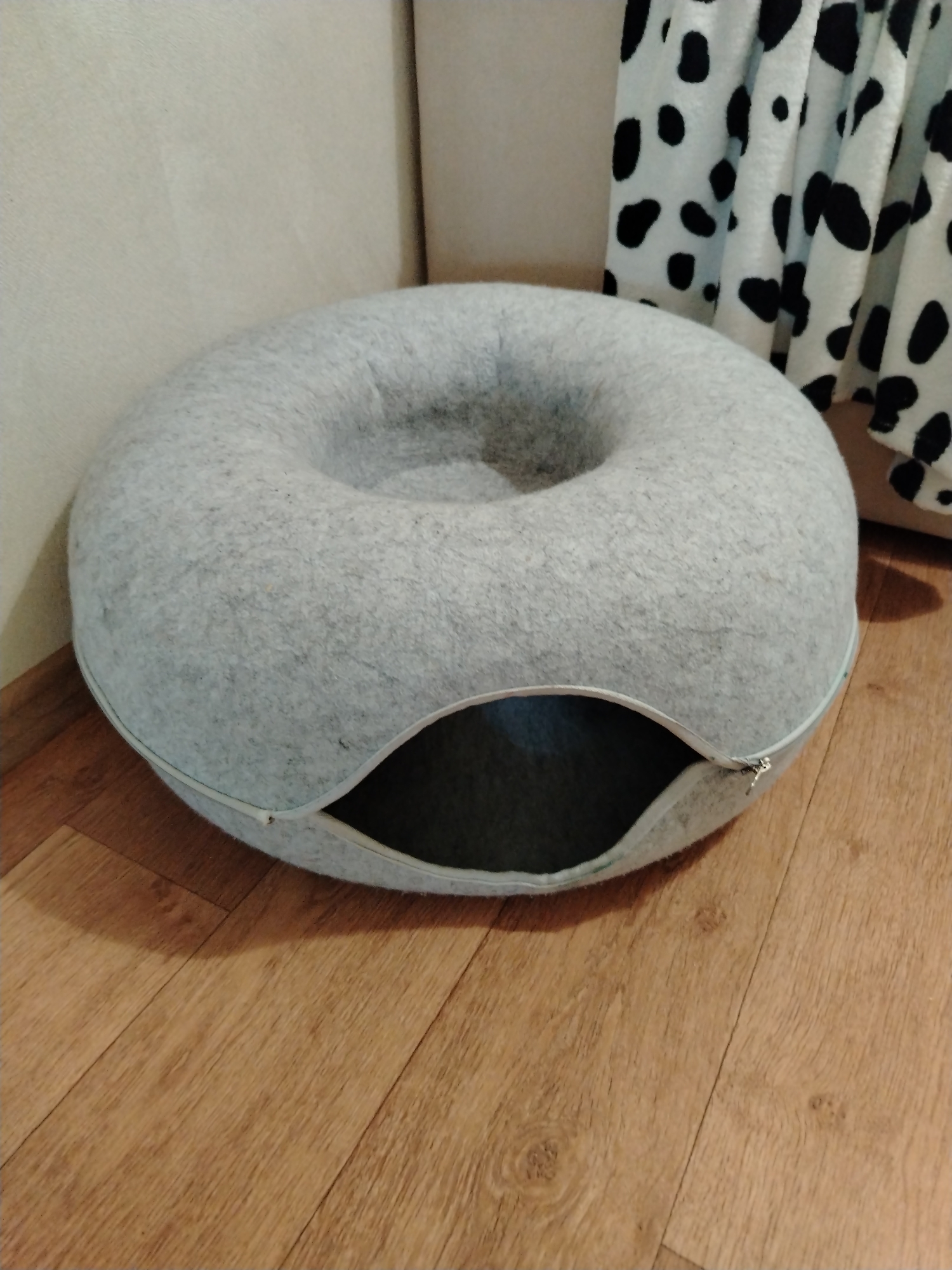An indoor scene displays a toroidal (donut-shaped) pet bed, predominantly light grey in color and made of a coarse, felt-like material, positioned centrally on a light brown wooden laminate floor. The pet bed features a circular depression in its upper surface and is constructed from two joined halves, indicated by a visible white zipper running along its lower circumference. A portion of this zipper on the front-right side is unzipped, creating a horizontal, crescent-shaped opening that reveals a dark interior cavity. The zipper pull tab is silver-colored.
To the left of the pet bed, a plain, pale beige or off-white wall is visible. To the right, a portion of a light-colored upholstered furniture piece (likely a sofa or bed) is present, draped with a white plush throw blanket adorned with irregular black spots, resembling a dalmatian pattern. The wooden laminate flooring consists of planks exhibiting varying wood grain patterns and subtle color differences, laid horizontally across the frame. No individuals or animals are visible. There is no discernible visual information within the image to suggest its specific location in the city of Смалявічы, Belarus.