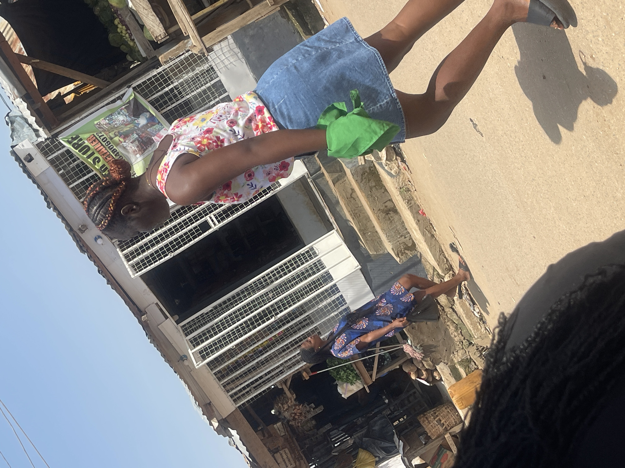 In Abuja, Nigeria, a street scene unfolds under a bright, clear sky. A woman, her hair styled in intricate braids with hints of orange, walks away from the viewer, her floral-patterned tank top and denim skirt creating a casual silhouette. In her left hand, she carries a folded bright green cloth. Another woman, further back and to the left, stands on a set of concrete steps, facing the first woman and looking away from the viewer, wearing a blue dress with a floral print. She holds a dark bag in her right hand. Behind them, a market stall with a metal grate facade displays various goods, including produce like cabbages and onions on a wooden table. A sign with text and an image hangs prominently, advertising a store. The ground is paved with asphalt, and sunlight casts long shadows. The environment suggests a bustling commercial area with a mix of permanent structures and temporary market stalls.