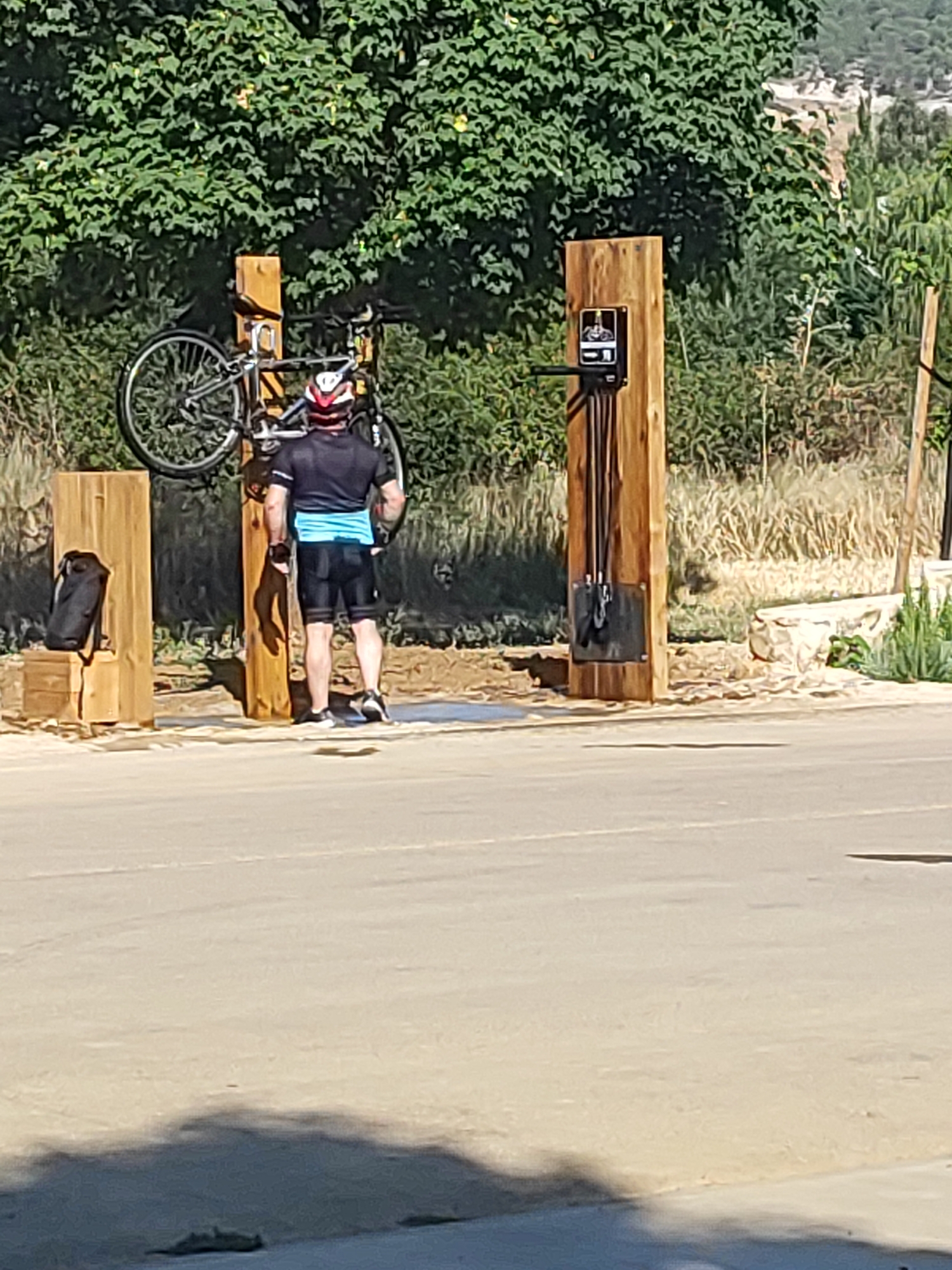 A male cyclist wearing a helmet, black and blue cycling jersey, and black cycling shorts stands with his bicycle suspended from a wooden post. The bicycle is hung by its frame, with the wheels facing outward. To the left of the cyclist, another wooden post has a black bag placed on it. To the cyclist's right, a third wooden post is equipped with a bicycle repair station, featuring a pump and various tools. The scene is outdoors, with lush green trees forming a backdrop, suggesting a park or recreational area. The ground is paved and sandy, with some dry vegetation visible behind the wooden posts. The lighting indicates it is daytime, with shadows cast by the objects. The overall setting is consistent with a designated rest or repair stop for cyclists in a natural environment, potentially within or on the outskirts of Villamuriel de Cerrato, Spain.