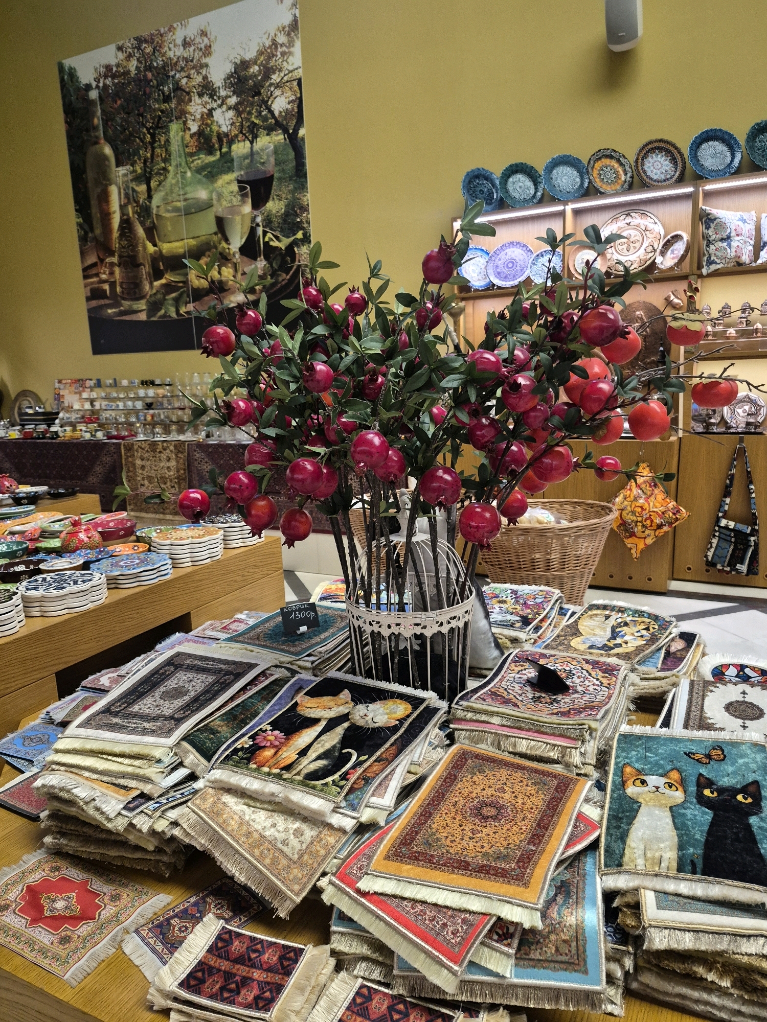 The image depicts a display of various decorative items, likely in a shop or market. In the foreground, numerous small rugs or carpets are stacked and spread out on a wooden surface. These rugs feature intricate patterns, some with traditional designs, while others display images of cats and celestial bodies. Behind the rugs, a decorative tree with artificial red pomegranate fruits is prominently displayed in a white lattice-like vase.
Further back, a large framed artwork hangs on a yellow wall, depicting a still life of wine bottles and glasses. To the right of the artwork, shelves are filled with decorative plates. Additional shelves in the background hold more pottery and other small decorative objects. A woven basket is visible behind the pomegranate tree.
The overall environment suggests a retail setting focused on decorative home goods and crafts. There are no people visible in the image. The style of the items, particularly the rugs and plates, hints at a possible Middle Eastern or Central Asian influence.