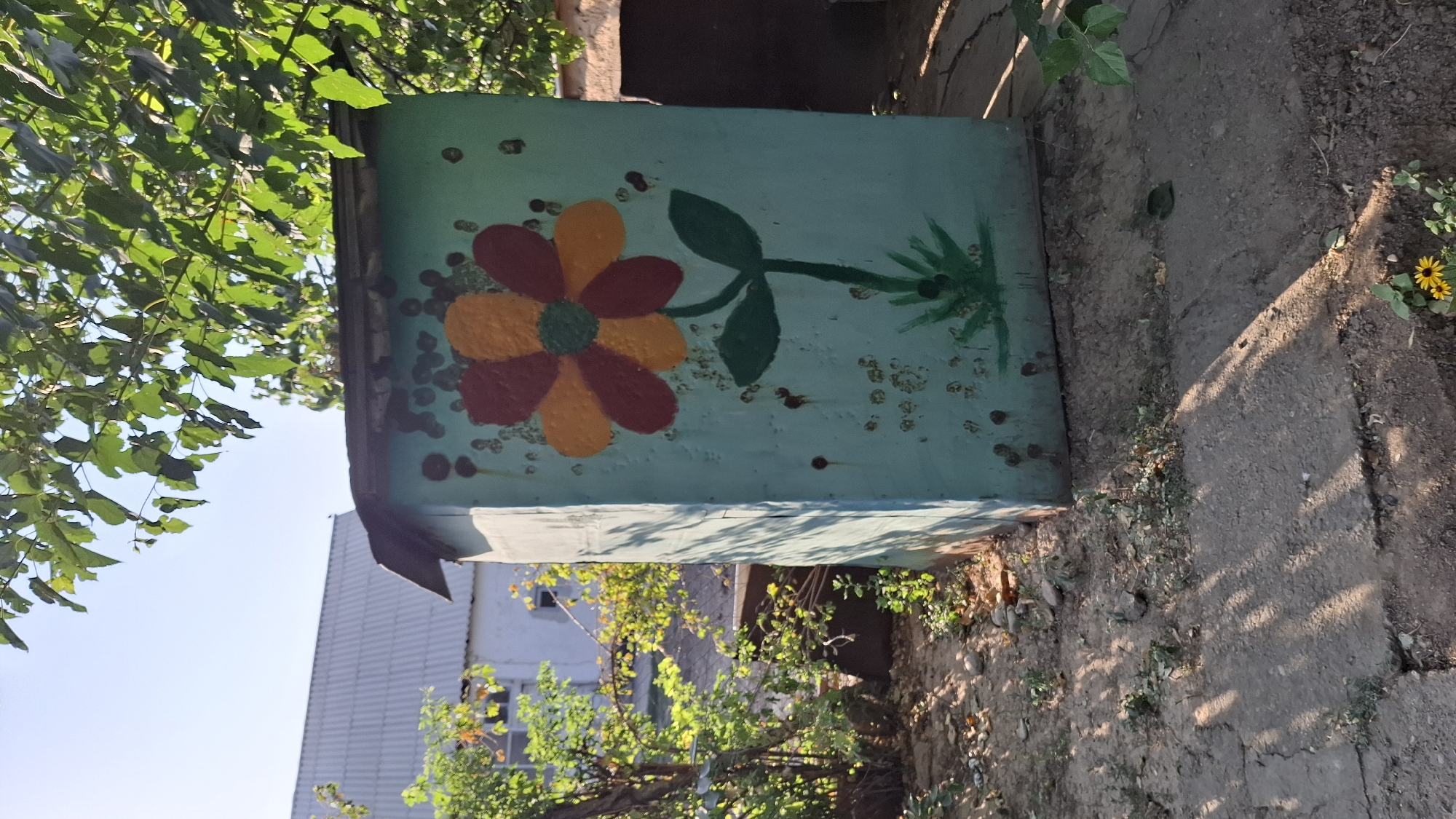 The image depicts an outdoor scene, likely in a rural or semi-rural area, with a structure that appears to be a small outhouse or shed in the foreground. The structure is painted a light blue-green and features a large, stylized painting of a flower with red and orange petals and a green center. The flower is depicted with a green stem and two leaves. The surface of the structure is weathered and shows signs of rust and minor damage. The ground surrounding the structure is uneven, with dirt, rocks, and patches of dry grass. Concrete slabs are visible in the foreground, showing cracks. In the background, a residential building with a corrugated metal roof can be seen, along with trees and foliage. Sunlight casts shadows across the ground, suggesting it is daytime. The overall impression is one of a rustic, lived-in environment. There are no people or animals visible in the image. The location is stated to be Шұбарсу, Kazakhstan.