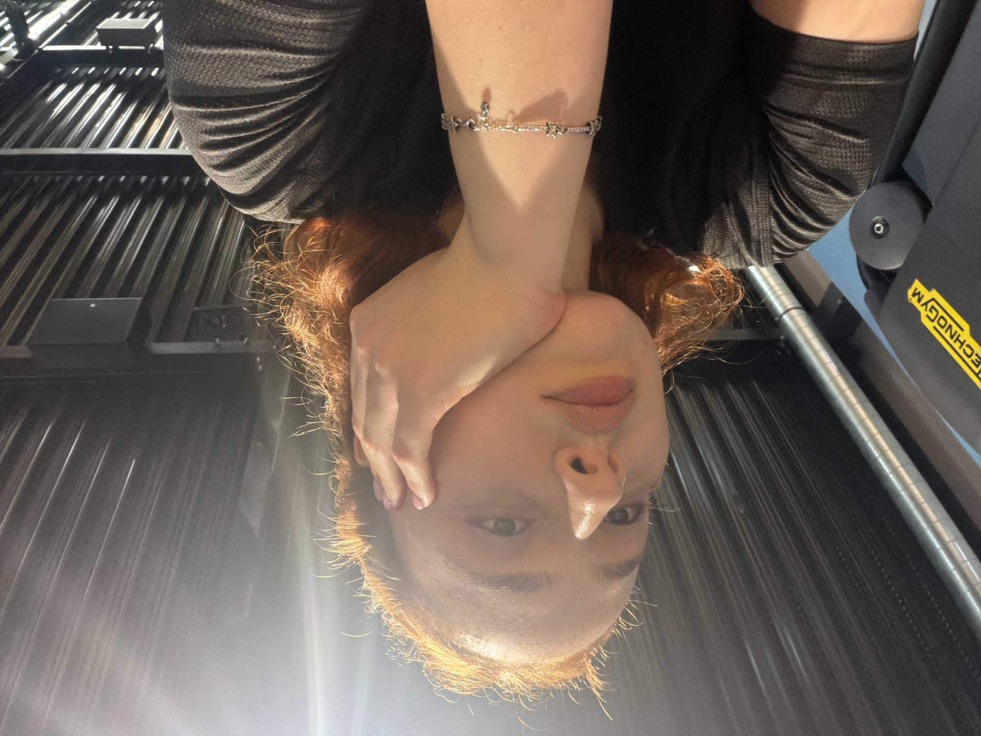 The image depicts a young adult female, captured in a close-up, looking directly at the viewer. She has reddish-brown hair, subtly illuminated, and is wearing a dark, long-sleeved top with a textured fabric. Her right hand, with light pink polished nails, is gently resting on her face, covering her chin and part of her cheek. A silver bracelet, adorned with various small charms including what appear to be stars and dark elements, is visible on her right wrist.
The setting is an indoor environment, likely a gym or fitness center. To the right of the person, a piece of dark gym equipment is visible, bearing the brand name "TECHNOGYM" in prominent yellow lettering. The background consists of dark, reflective, possibly metallic or corrugated panels, creating linear patterns from reflected light. Bright light sources cause glares on these background surfaces and highlight features on the person's face, such as her forehead and nose. The expression on her face is neutral, suggesting a posed shot. The location context provided is Strasbourg, France.